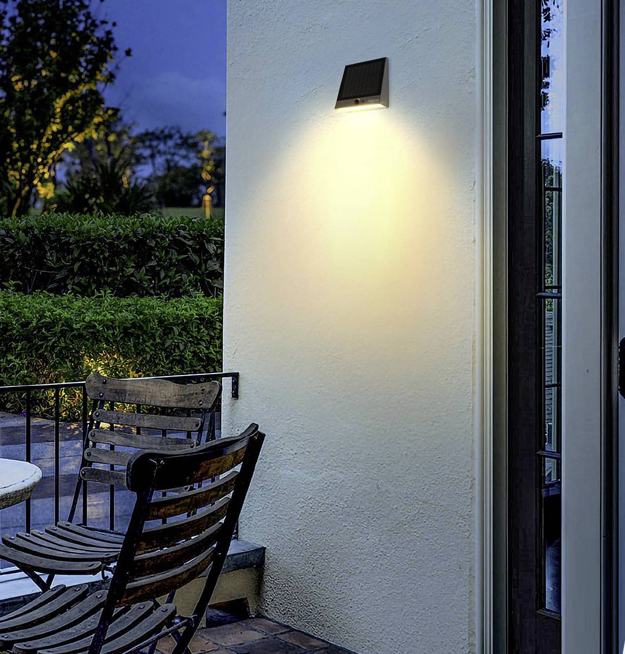 Night-time scene of a garden with wooden chairs on a terrace. A wall light illuminates part of the wall. Trees are visible in the background.