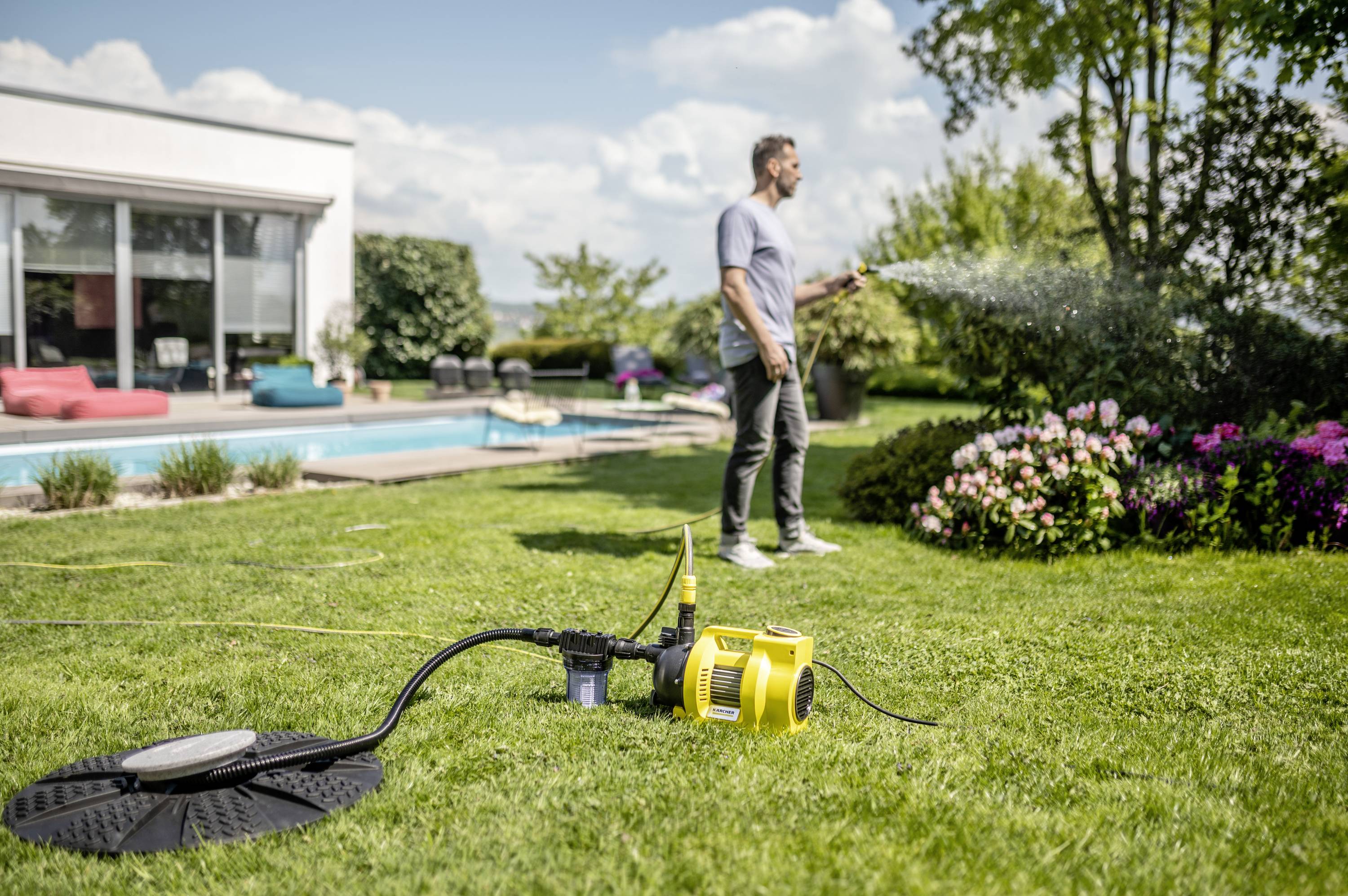 A man is watering the garden with a hosepipe. In the foreground stands a yellow pump. In the background is a house with a swimming pool.