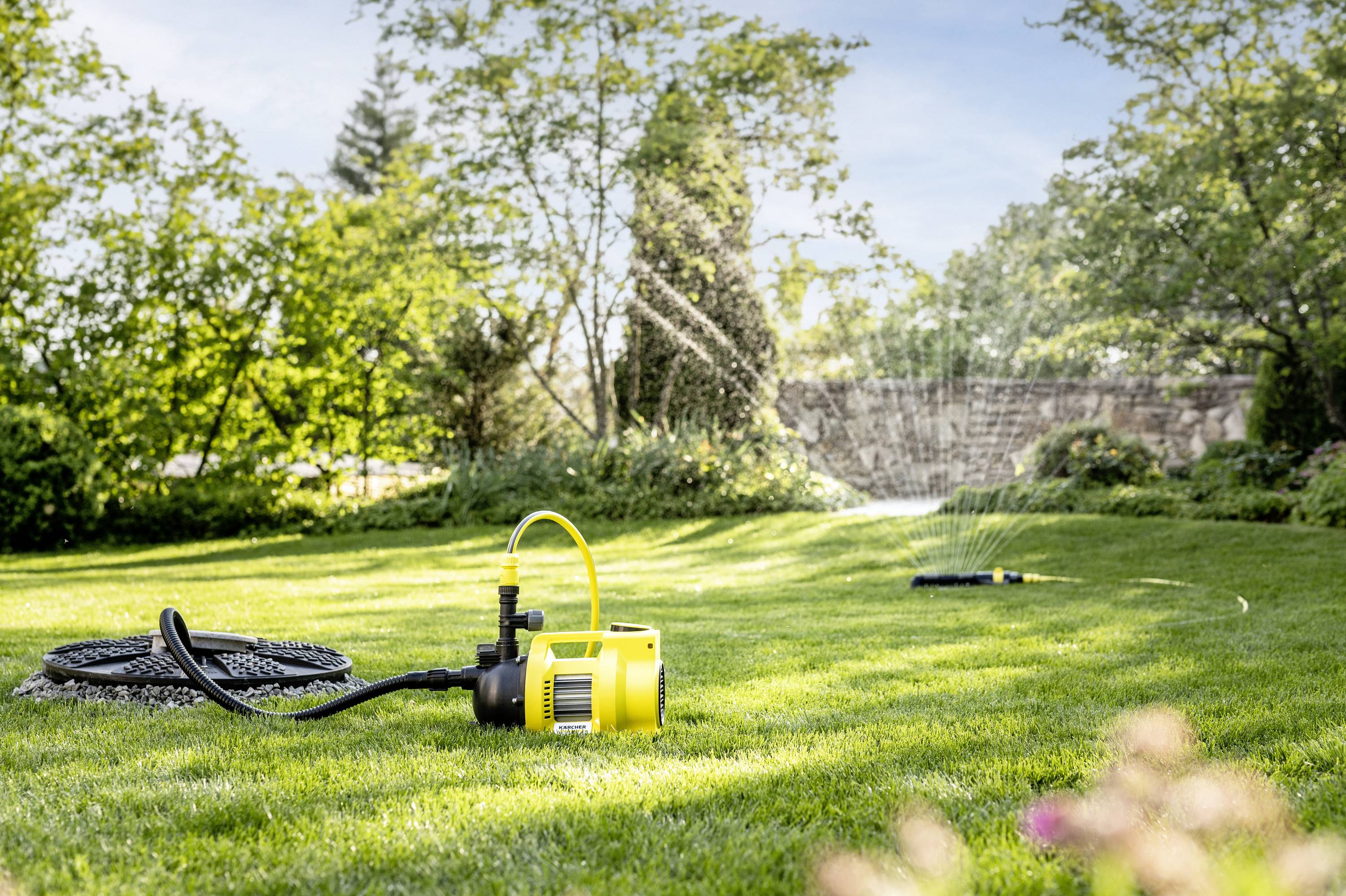 A yellow water pump stands on a green lawn. A sprinkler nearby is watering the garden. Trees can be seen in the background.