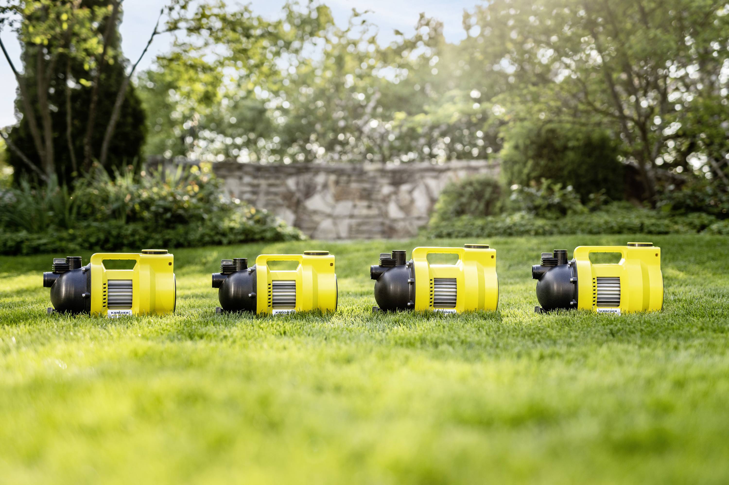 Four yellow water pumps stand side by side on green grass in the garden on a sunny day, surrounded by trees and bushes.