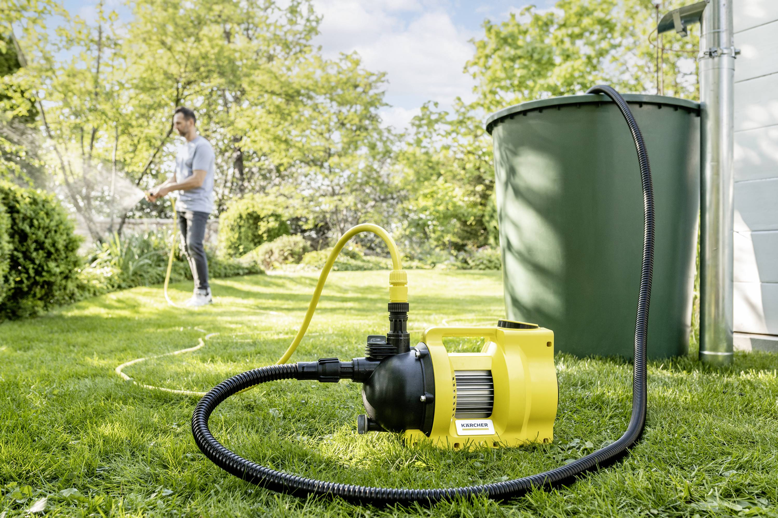 A yellow water pump on grass with an attached hose. In the background, a person is watering plants, with a green water barrel beside them.