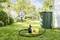 A man is watering the garden with a hosepipe. In the foreground, a yellow pump is connected to a rainwater tank.