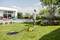 A man is watering the lawn with a garden hose next to a modern house with a swimming pool, surrounded by flower beds and trees.