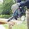A person is sawing a wooden board outdoors using an electric saw. Wood shavings are flying, and the surrounding area is green and wooded.