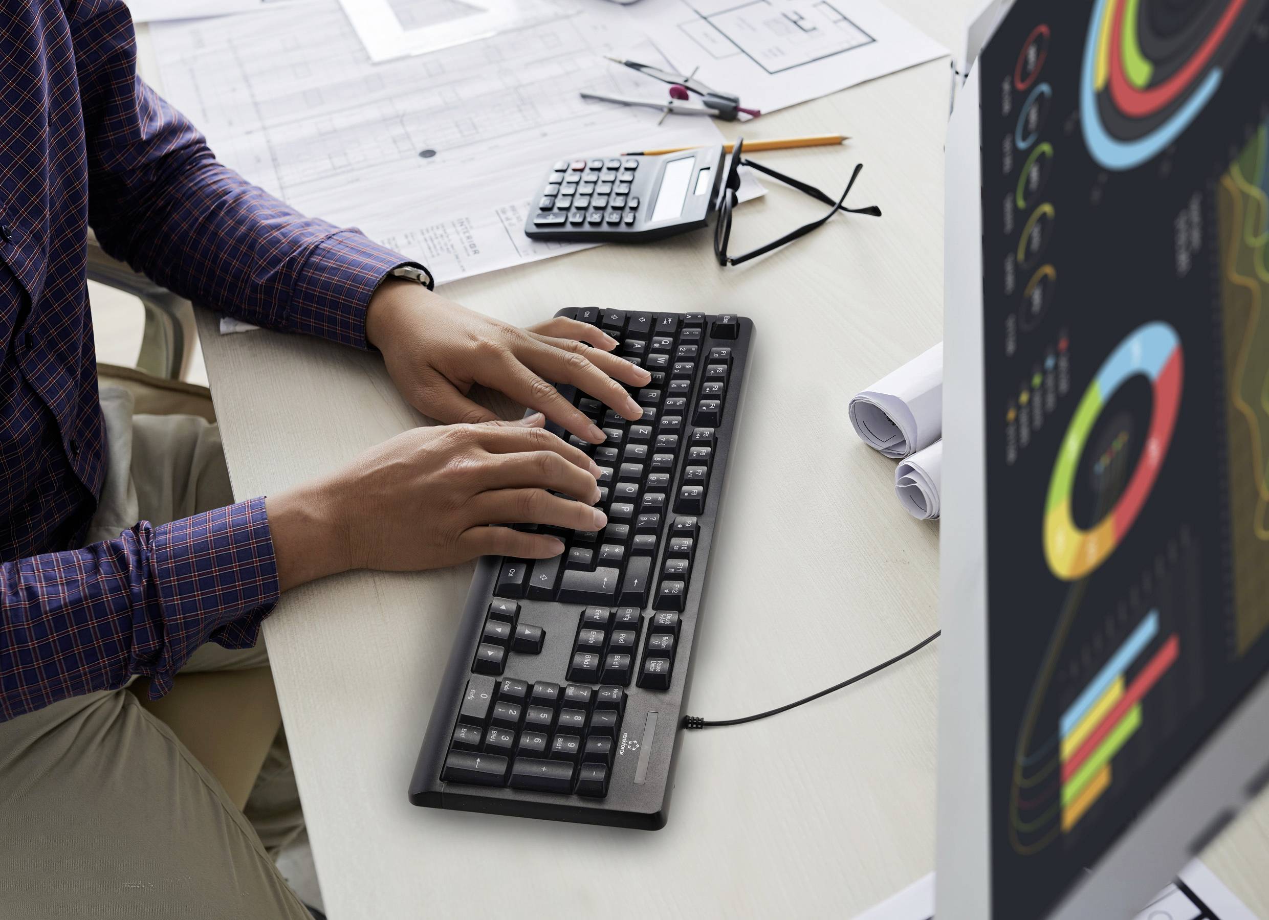 A person is typing on a keyboard at a desk with diagrams on a screen, papers, and a calculator.