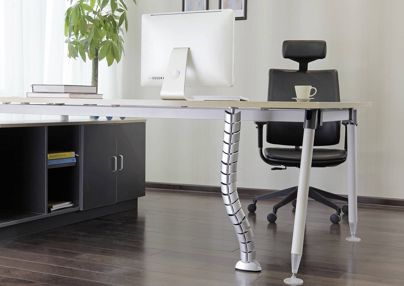 A modern office with a desk featuring a computer and a coffee mug. Beside it, an office chair with plants in the background.