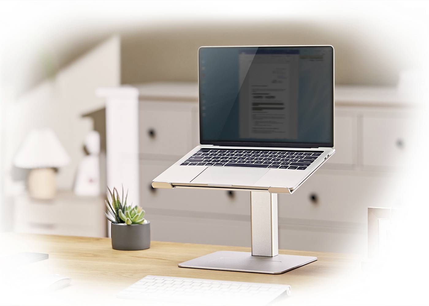A laptop sits on a desk stand in front of a blurred interior backdrop. A small cactus and a keyboard are visible.