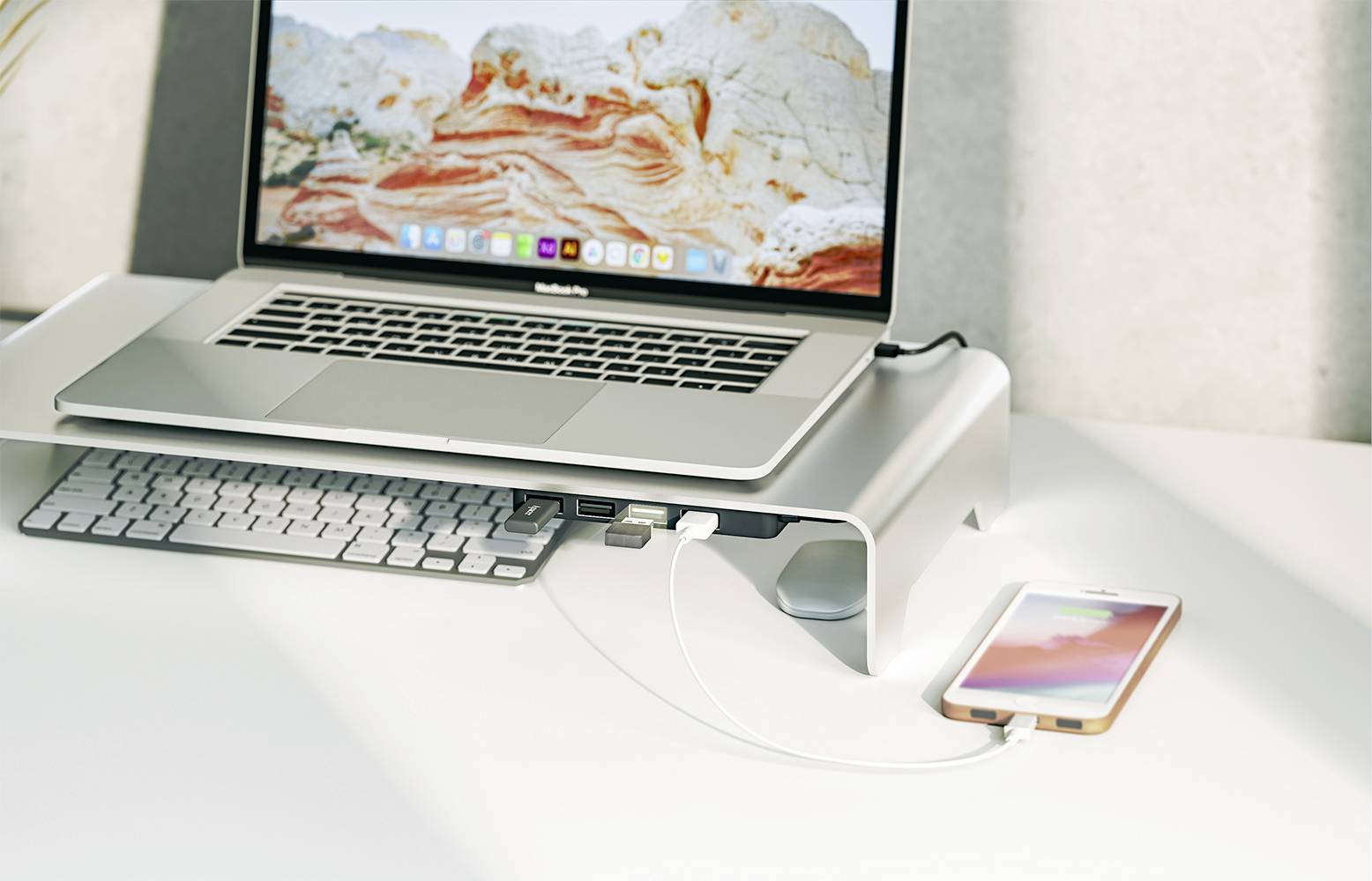 A laptop on a stand, connected to a smartphone. A keyboard beneath it. The screen displays a landscape.