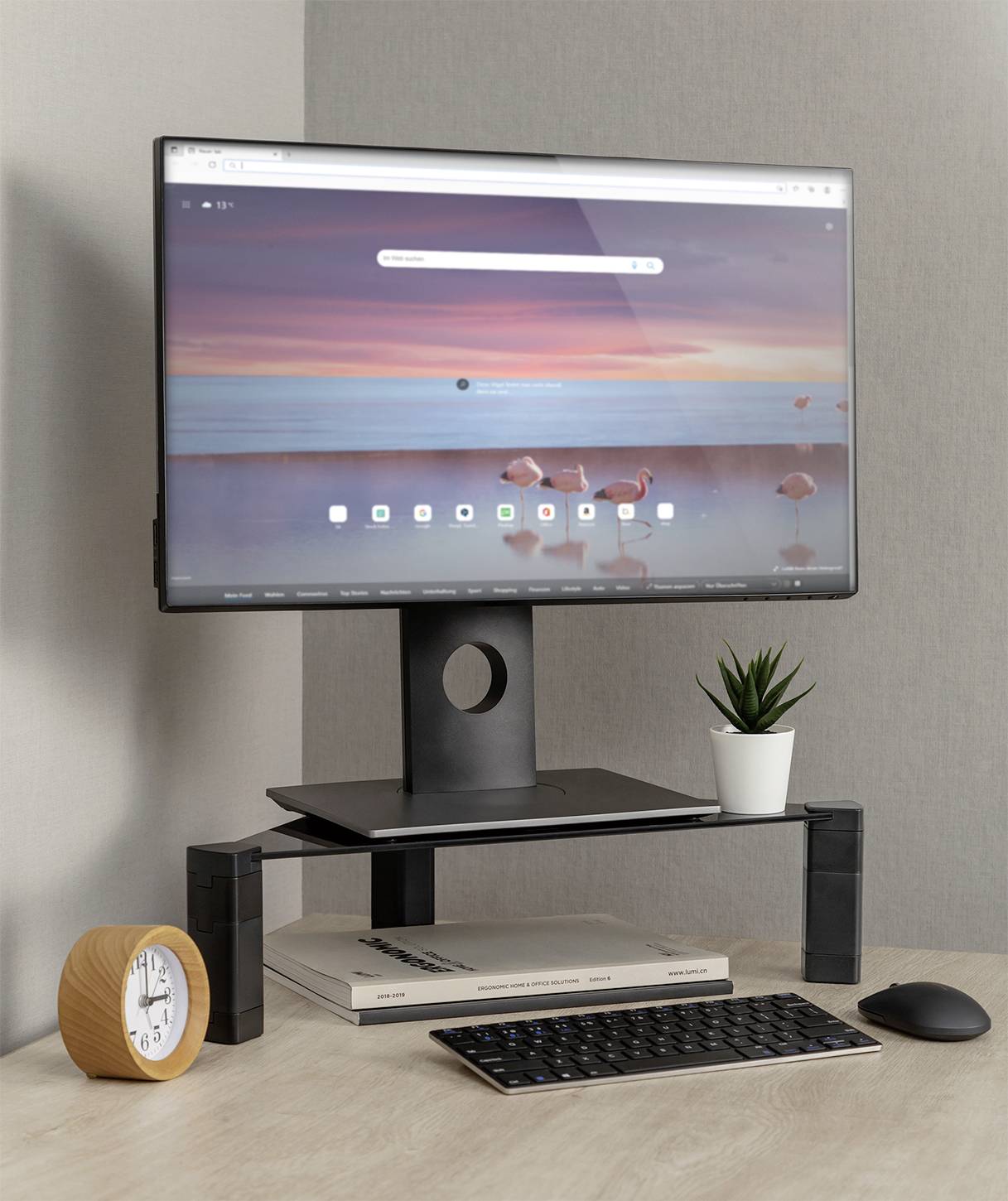 A computer screen displays a coastal scene. Beneath it, books, a small plant, a clock, a keyboard and a mouse sit on a desk.