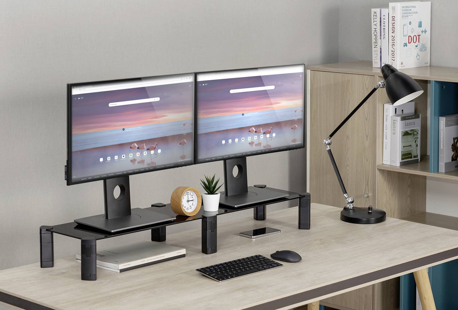 Two monitors on a desk, supported by a monitor stand. Beside them are a desk lamp, an alarm clock, and some plants.