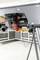 Underneath view of a table saw with dust extractor connection and power cable in a workshop with white shelves and grey floor.