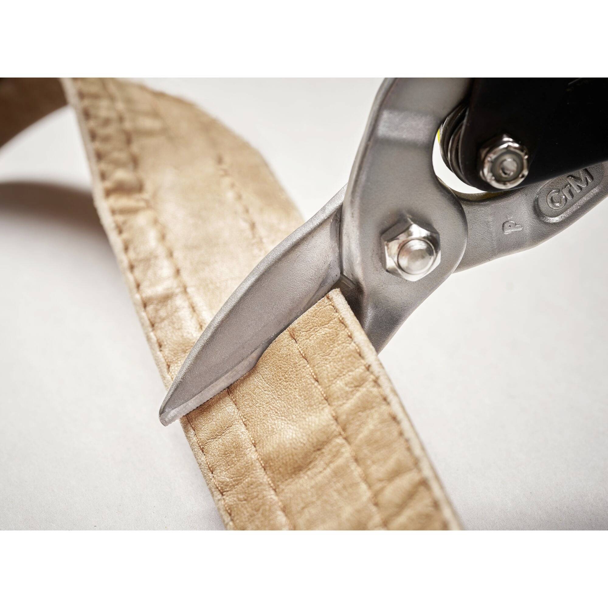 A pair of scissors cuts through a thick brown leather strap. Close-up reveals the cutting edge and the texture of the leather.