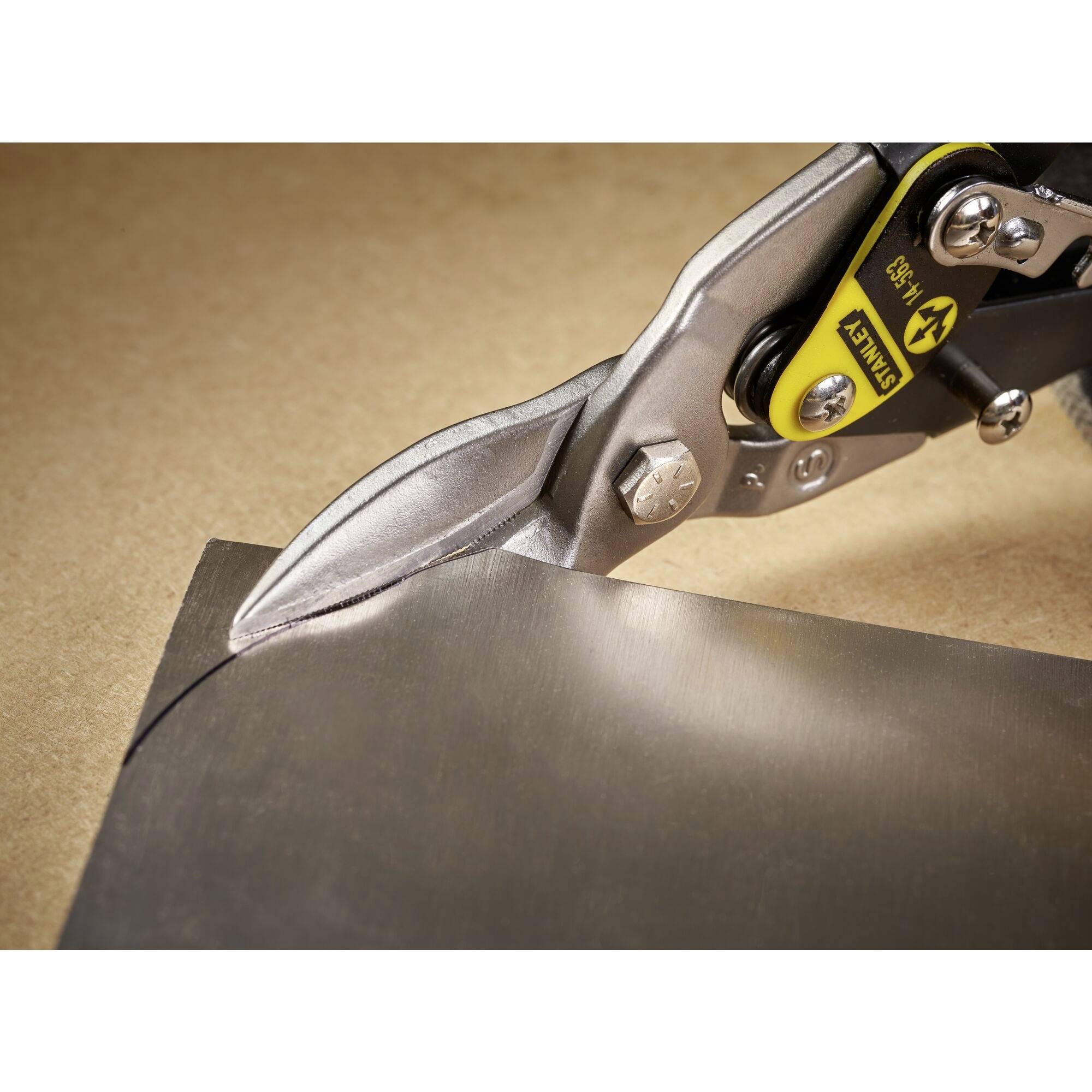 A metal sheet cutter slices through a thin sheet of metal on a wooden table. The blade is sharp and sturdy.