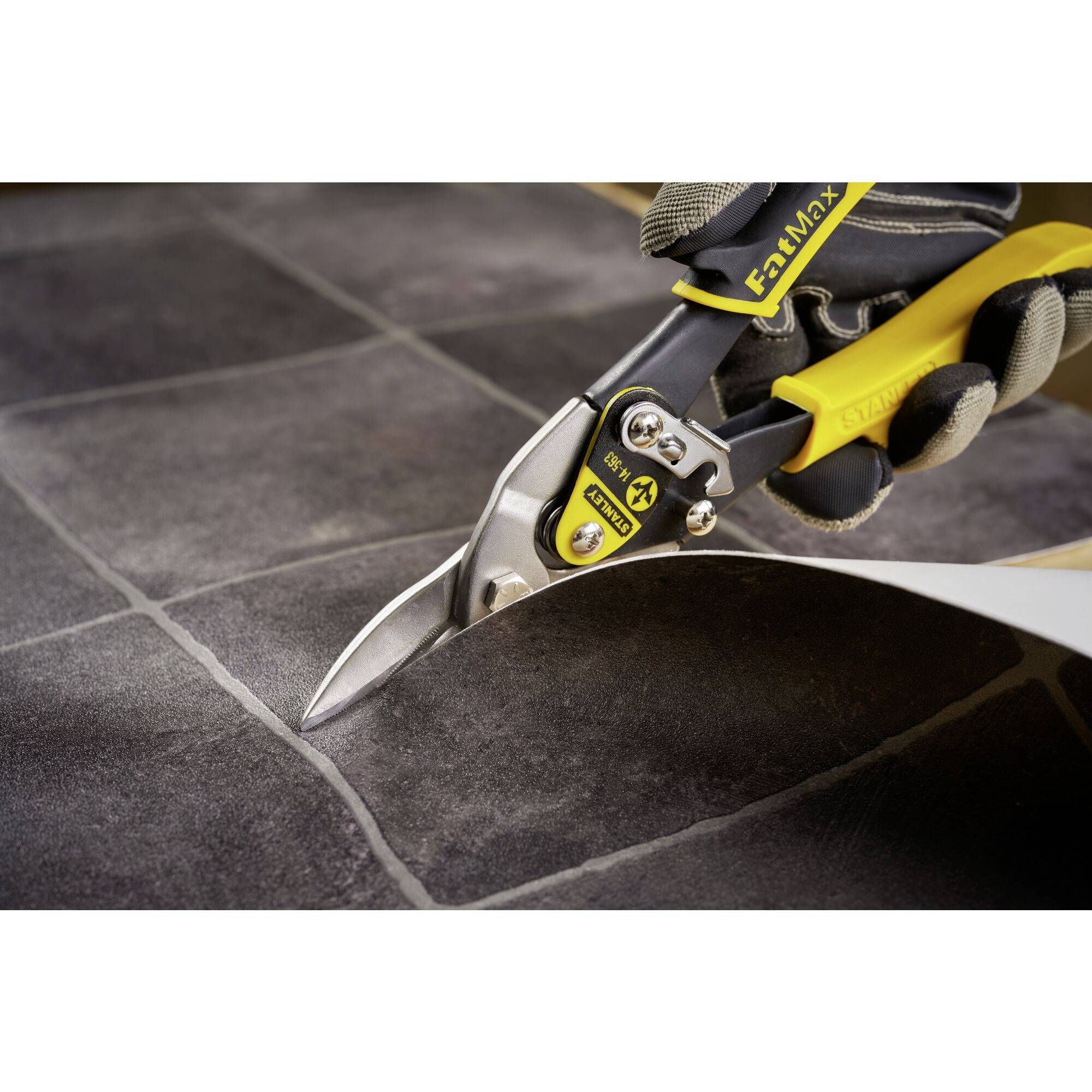 A person is cutting a piece of metal with yellow and black metal shears on grey tiles. The shears have ergonomic handles.