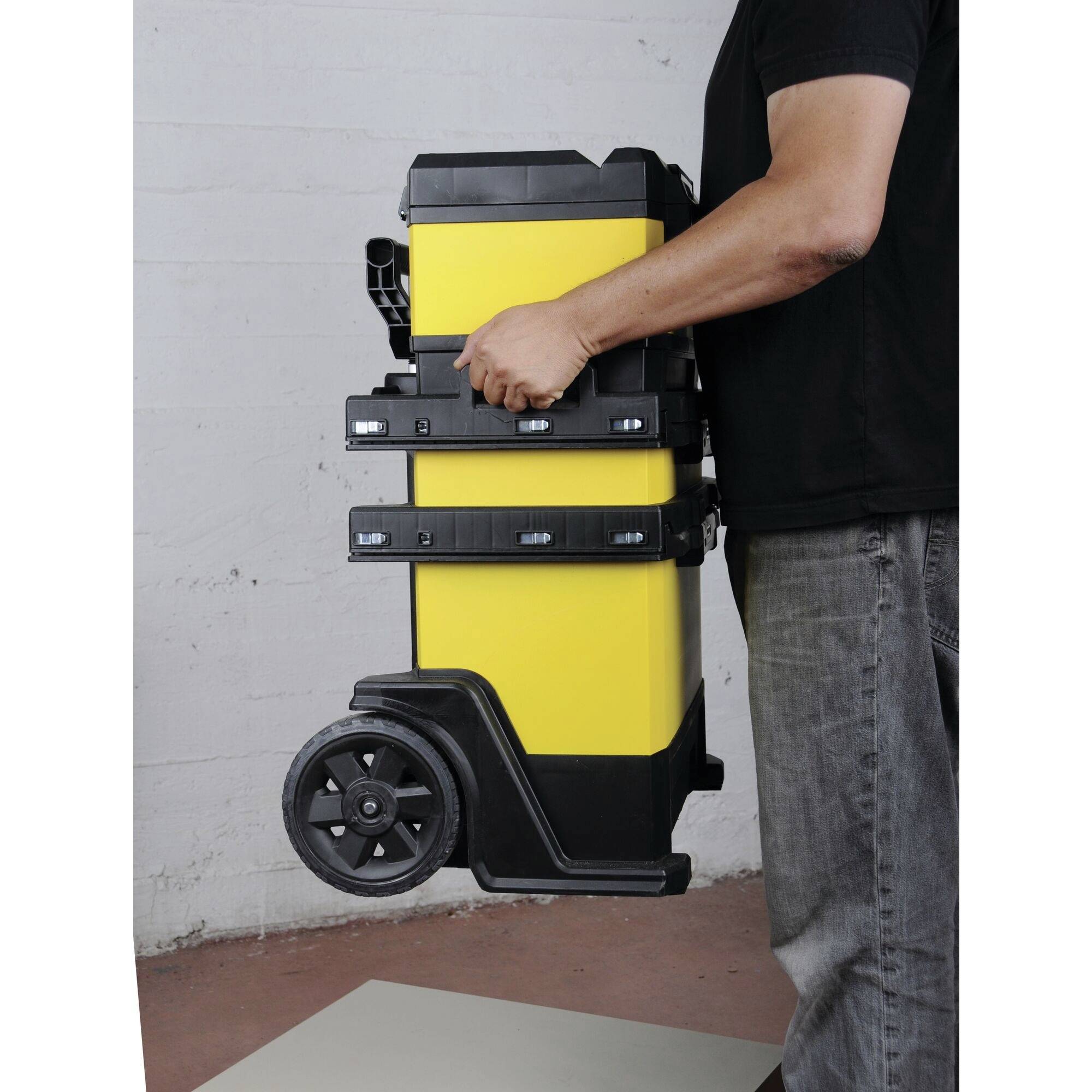 A person is transporting a yellow wheeled toolbox. The case has multiple stackable compartments and is carried at its side.