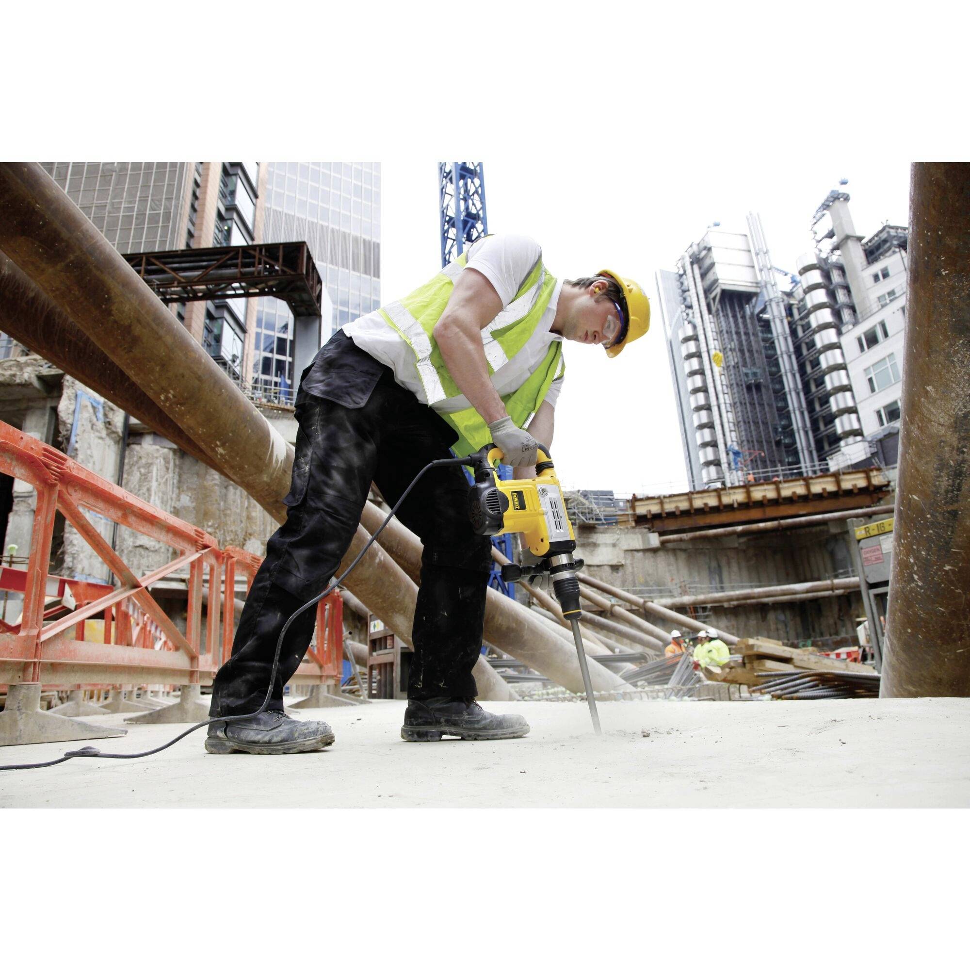 A construction worker wearing a high-visibility vest and hard hat is using a pneumatic drill on a building site in an urban setting with skyscrapers.