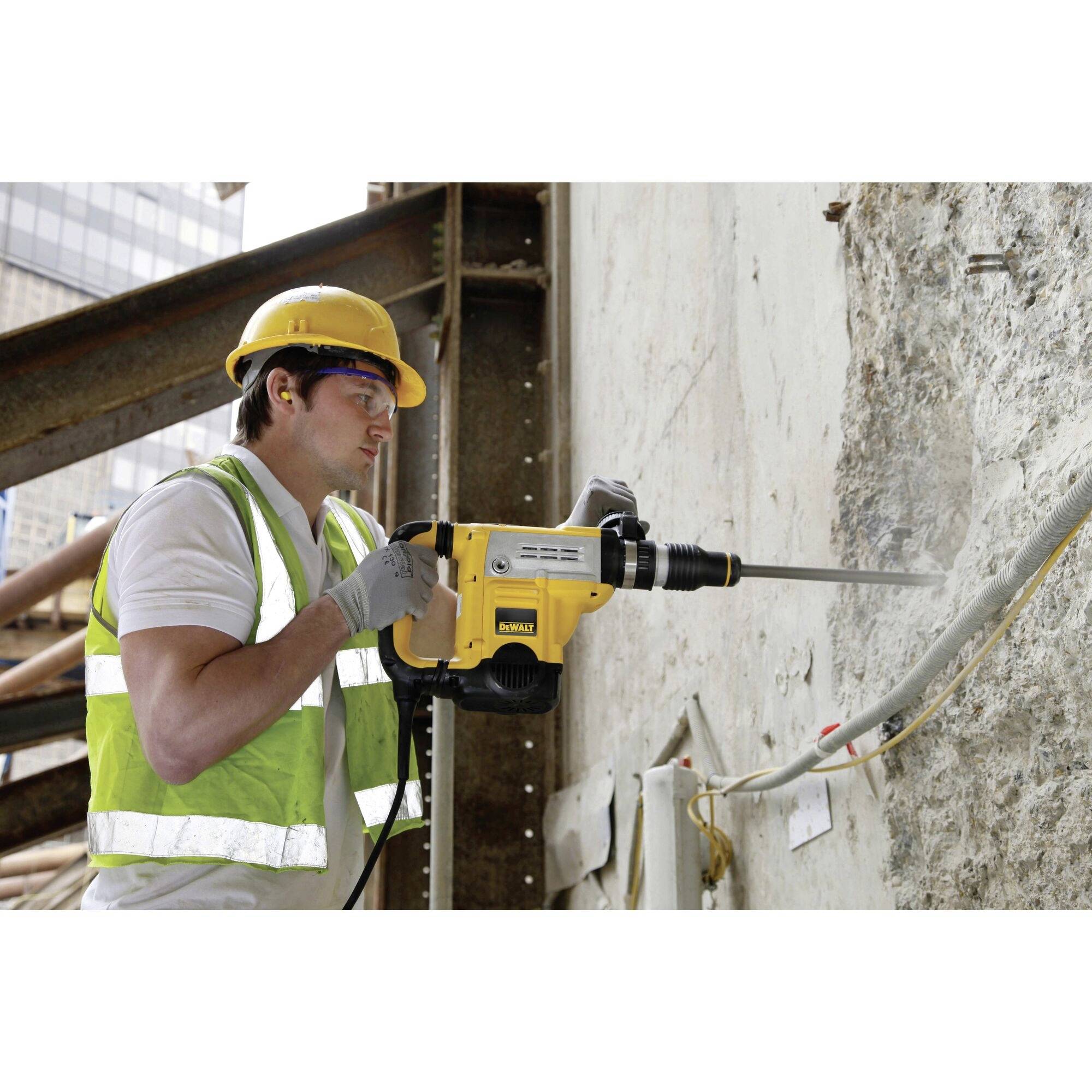 A person in safety equipment is using a yellow hammer drill to bore a hole in a concrete wall.