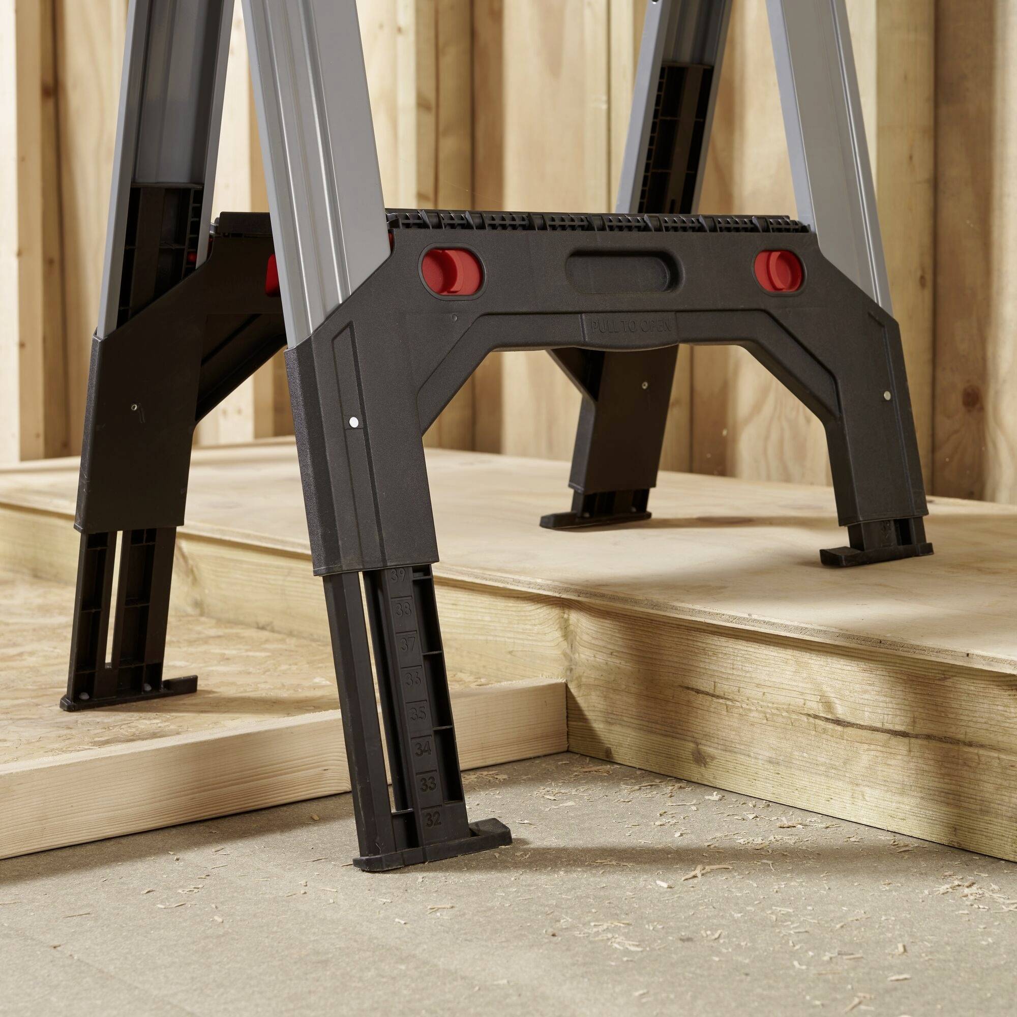 A foldable metal ladder in a workshop environment, set up on a wooden platform, with red locking buttons.