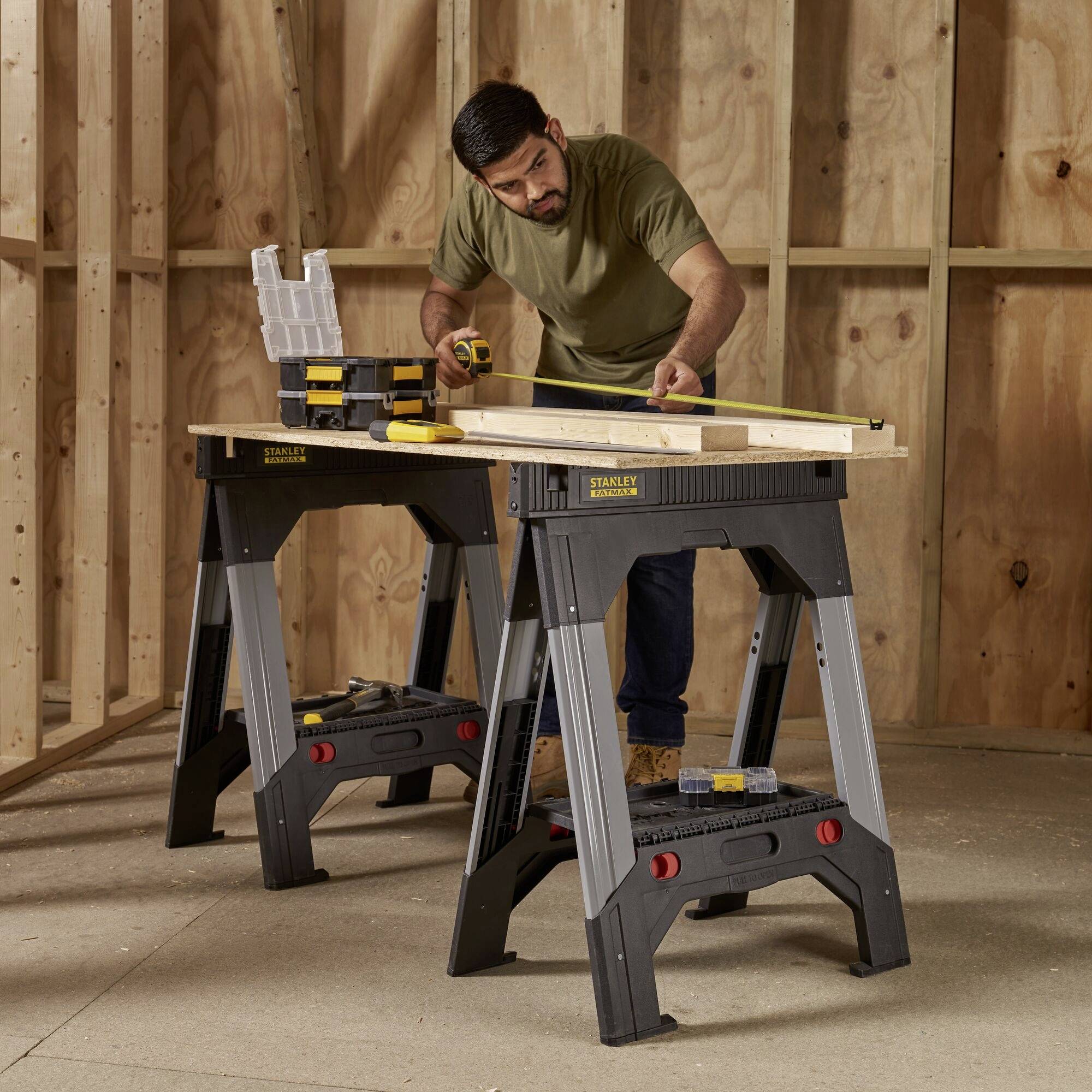 A man in workwear is measuring a wooden board on a workbench in a woodworking workshop.