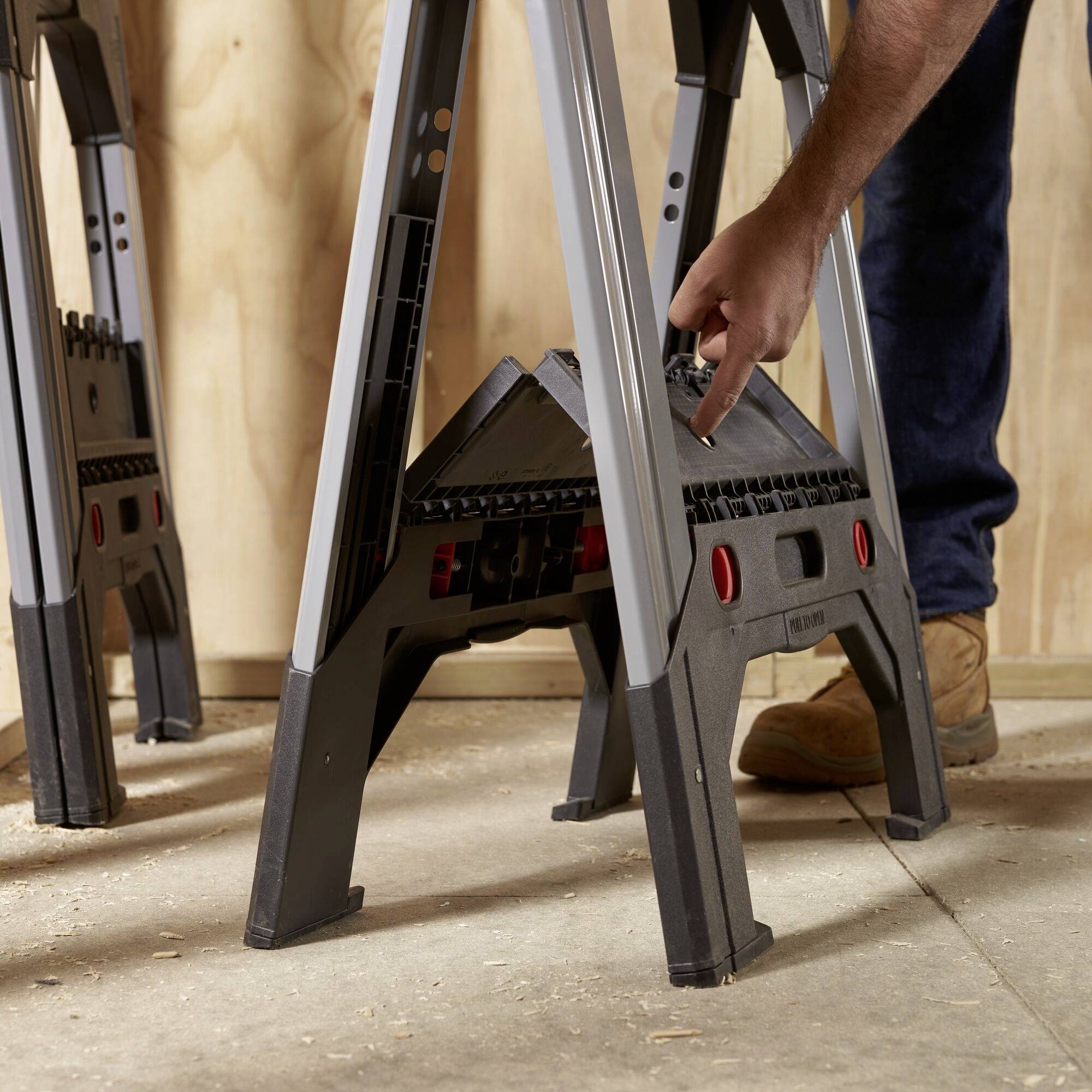 A person is positioning two folding sawhorses in a workshop. The floor is covered with wooden boards.