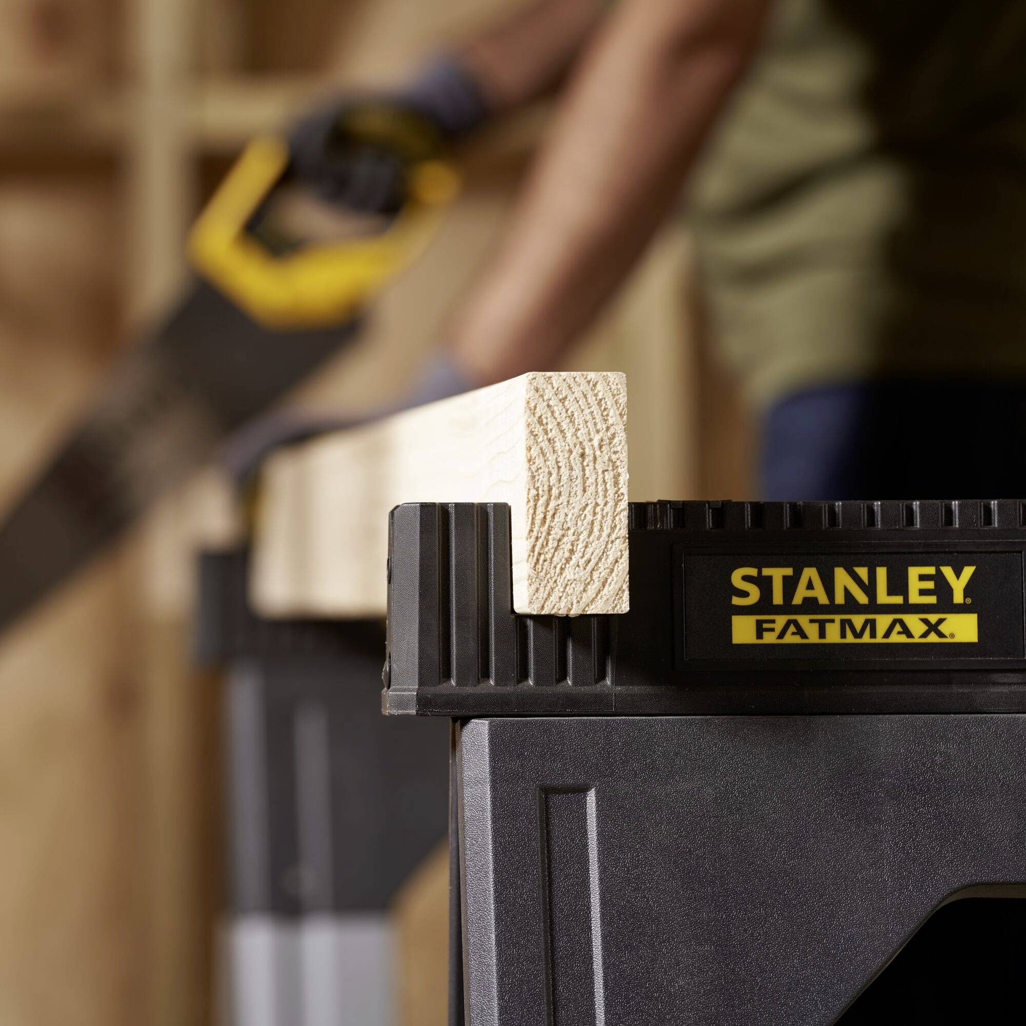A piece of wood lies on a Stanley FatMax sawbench. In the background, a blurred person can be seen holding a tool.