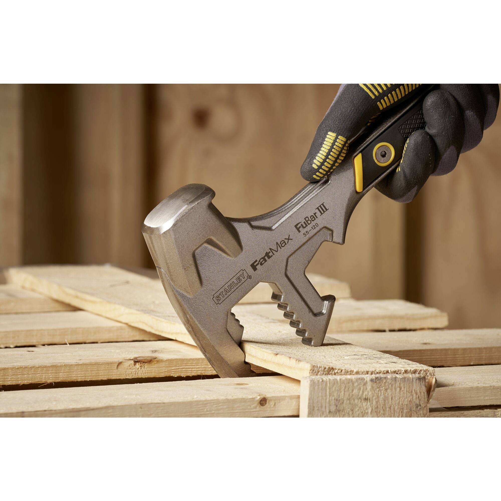 A sturdy hammer head is working on wood. A gloved hand grips the hammer firmly. Timber is stacked in the background.