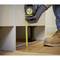 A hand wearing a glove holds a tape measure, measuring the height between the floor and the underside of a piece of furniture. The person is wearing work trousers and boots.