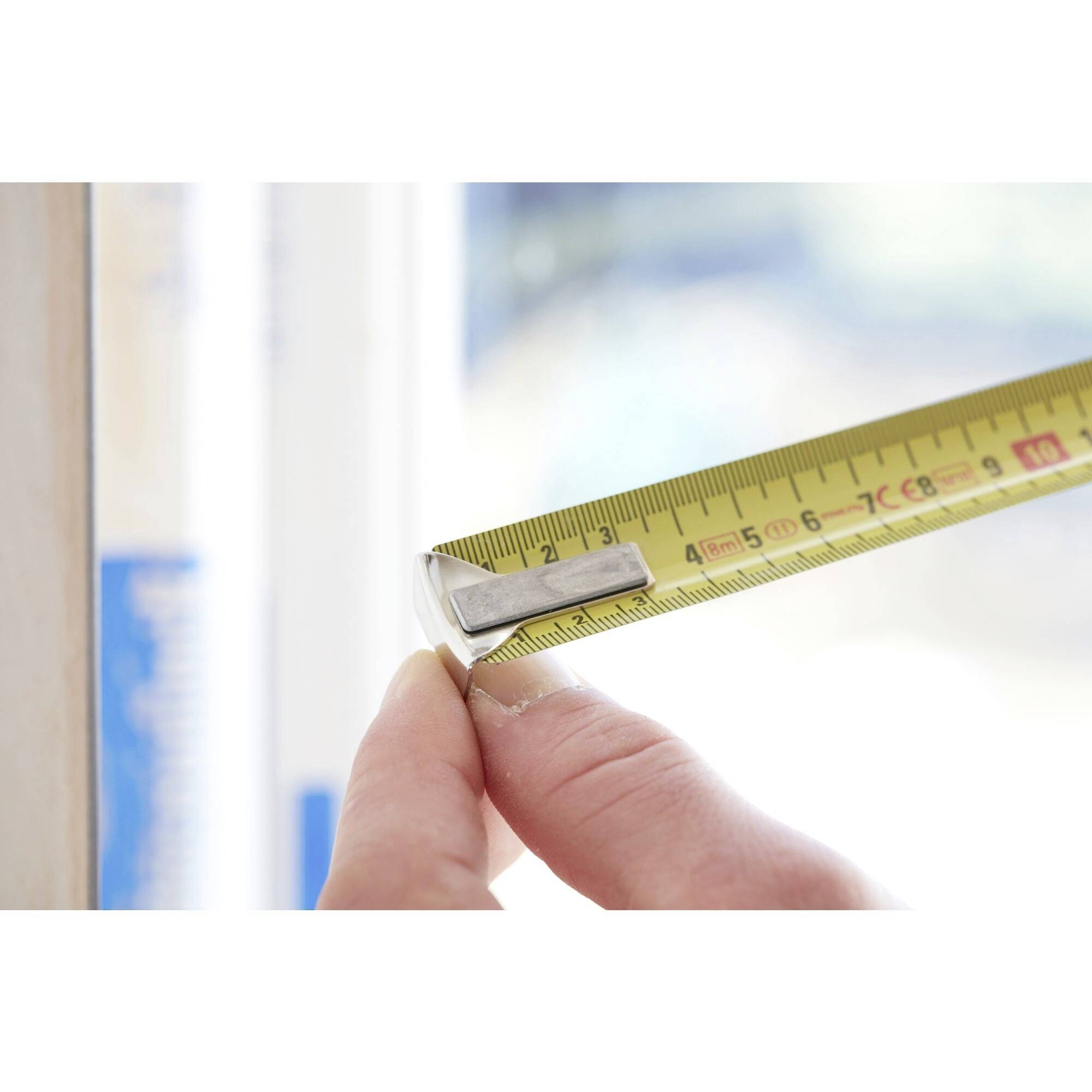 A hand holds a tape measure, measuring the edge of a window. The focus is on precise measurement and the tape measure itself.