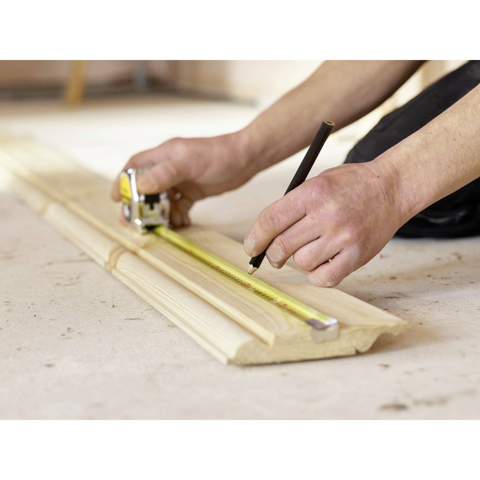 A person measures a wooden board with a tape measure and marks the spot with a pencil.