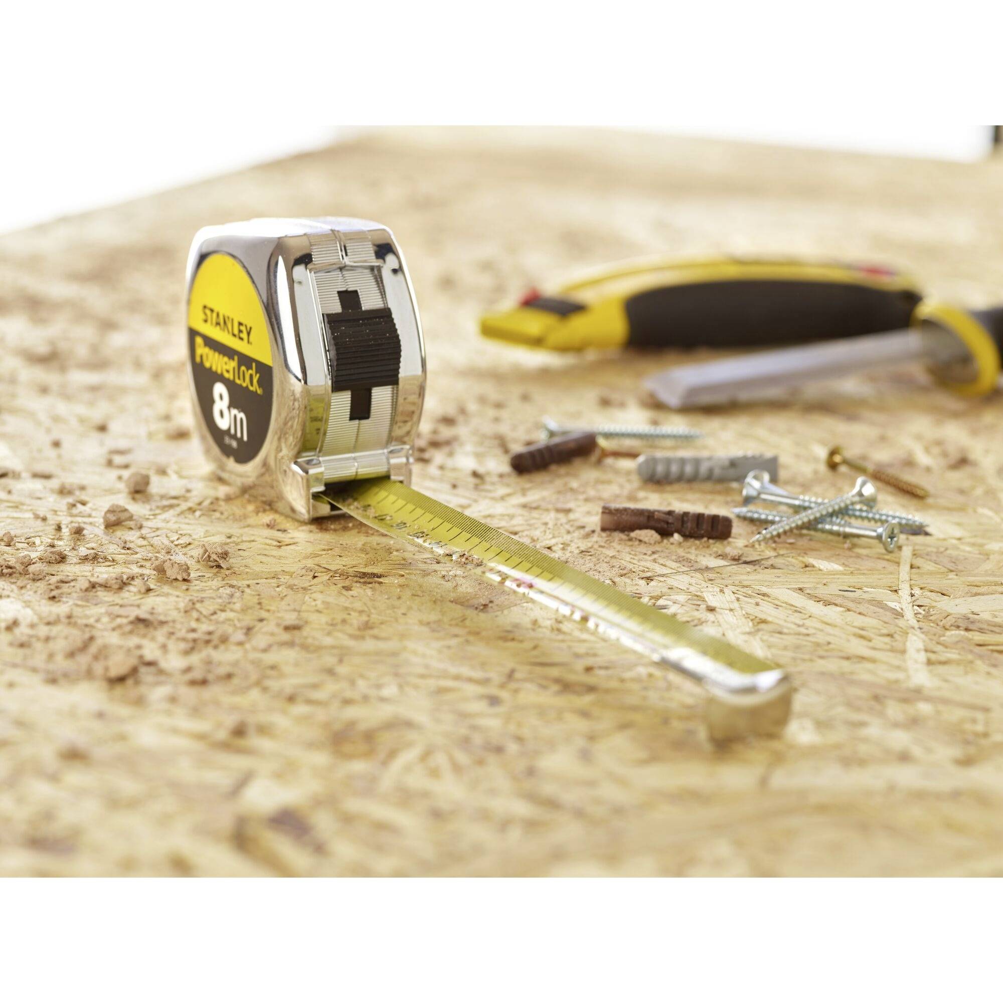 A measuring tape lying on a table with an 8-metre scale, surrounded by screws and a Stanley knife on a wooden surface.