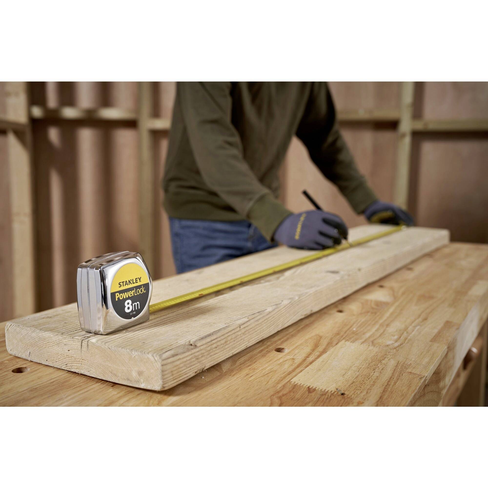 A person is measuring a wooden board with a tape measure on a table. They are wearing gloves and making markings with a pencil.