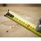 Close-up of a yellow tape measure lying on a wooden surface with wood shavings. A gloved hand is holding the end of the tape.
