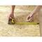 A person is measuring a piece of plywood with a yellow tape measure and marking it with a pencil for a precise cut.