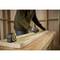 A person is measuring a wooden board with a tape measure in a workshop. On the table lies a 5m tape measure with a yellow casing.