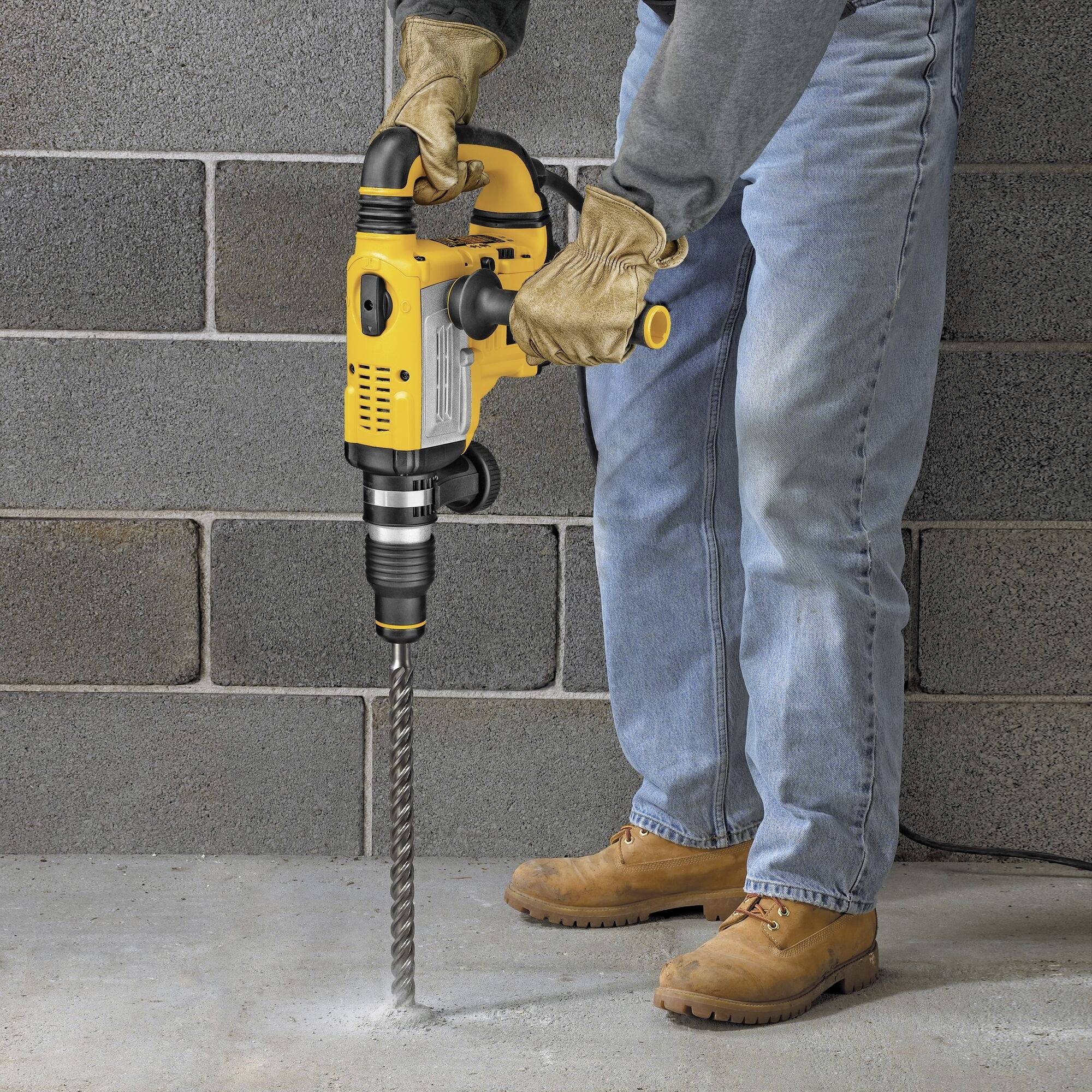 A person wearing jeans is drilling a hole in a concrete wall using a large yellow drill. They are wearing safety glasses and work gloves.