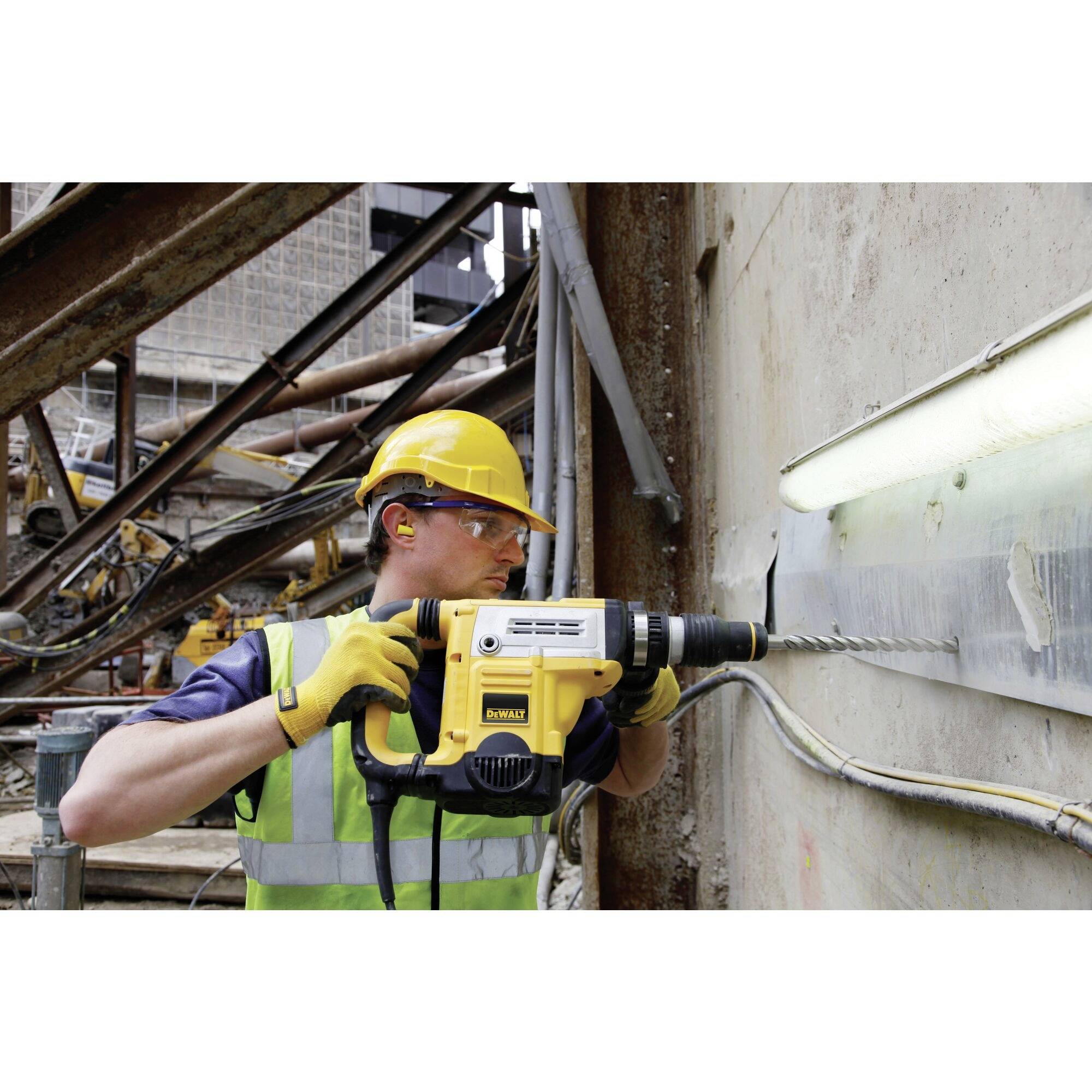 A construction worker wearing a hard hat and safety glasses is drilling into a concrete wall with a yellow drill on a building site.