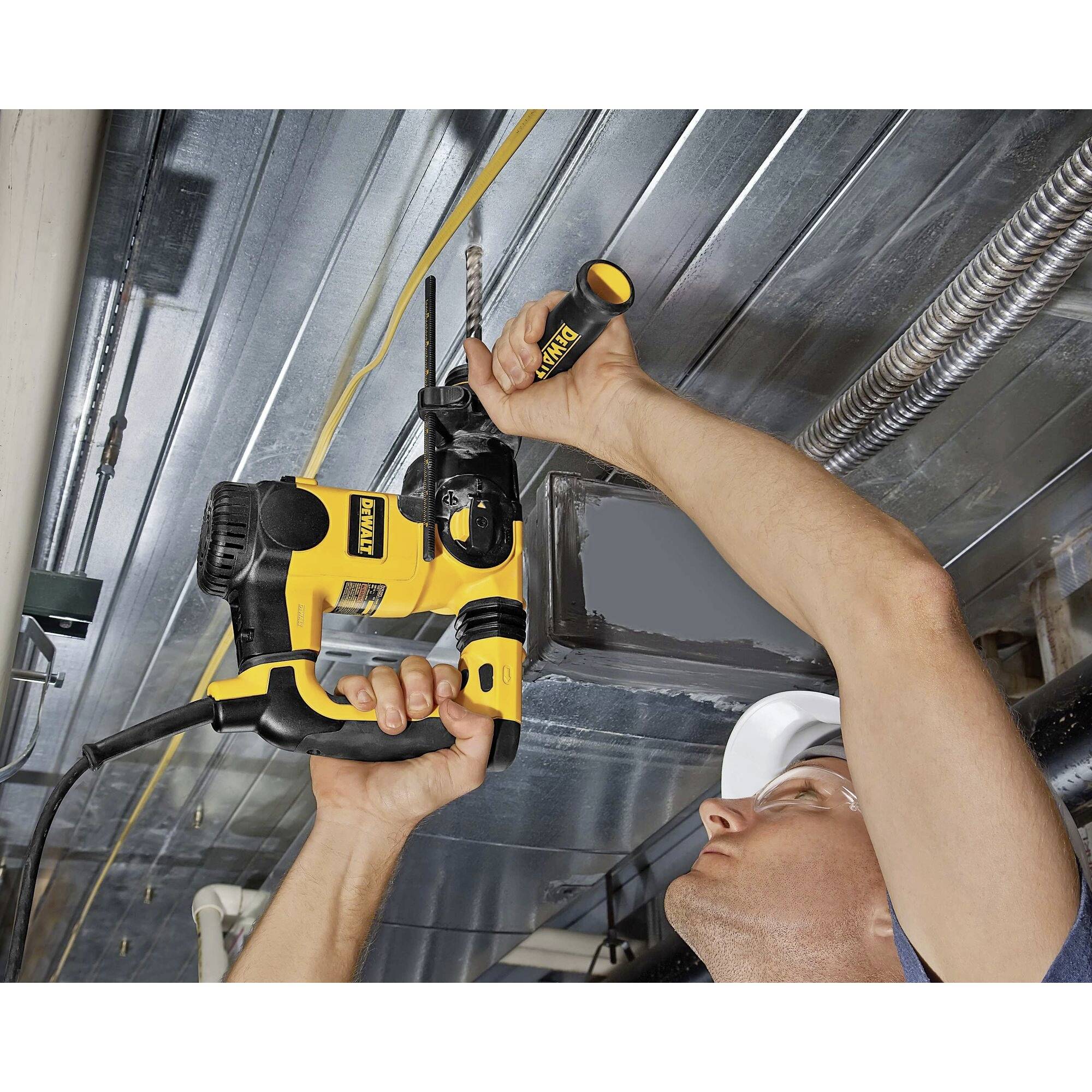 A person is drilling into the ceiling of a building with a yellow drill. Surrounded by pipes and tunnel conduits.