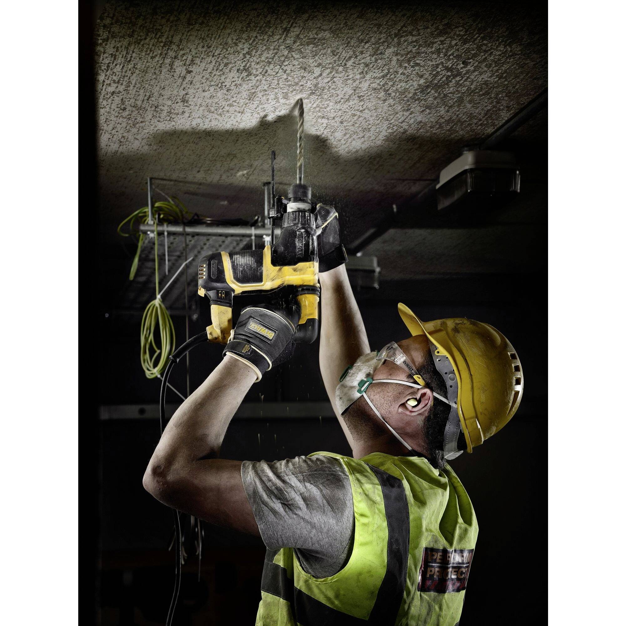 A construction worker in safety gear is drilling a hole in a ceiling with a yellow machine, wearing safety glasses and a hard hat.
