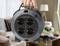 A hand holds a round black multi-socket cable reel plug in front of a living room background.