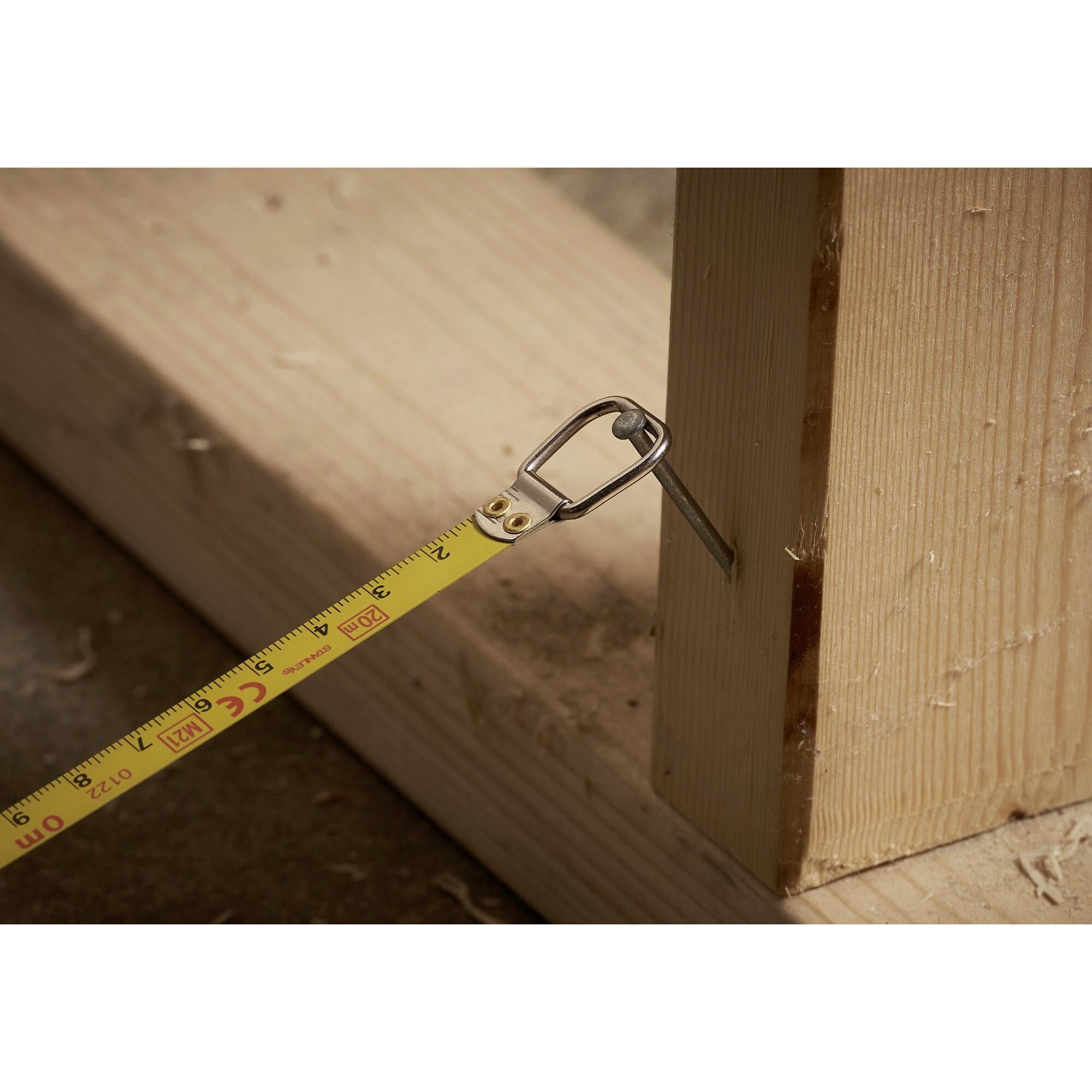 A tape measure is measuring the width of a wooden beam on a construction site. The tape is attached to a hook on the beam.