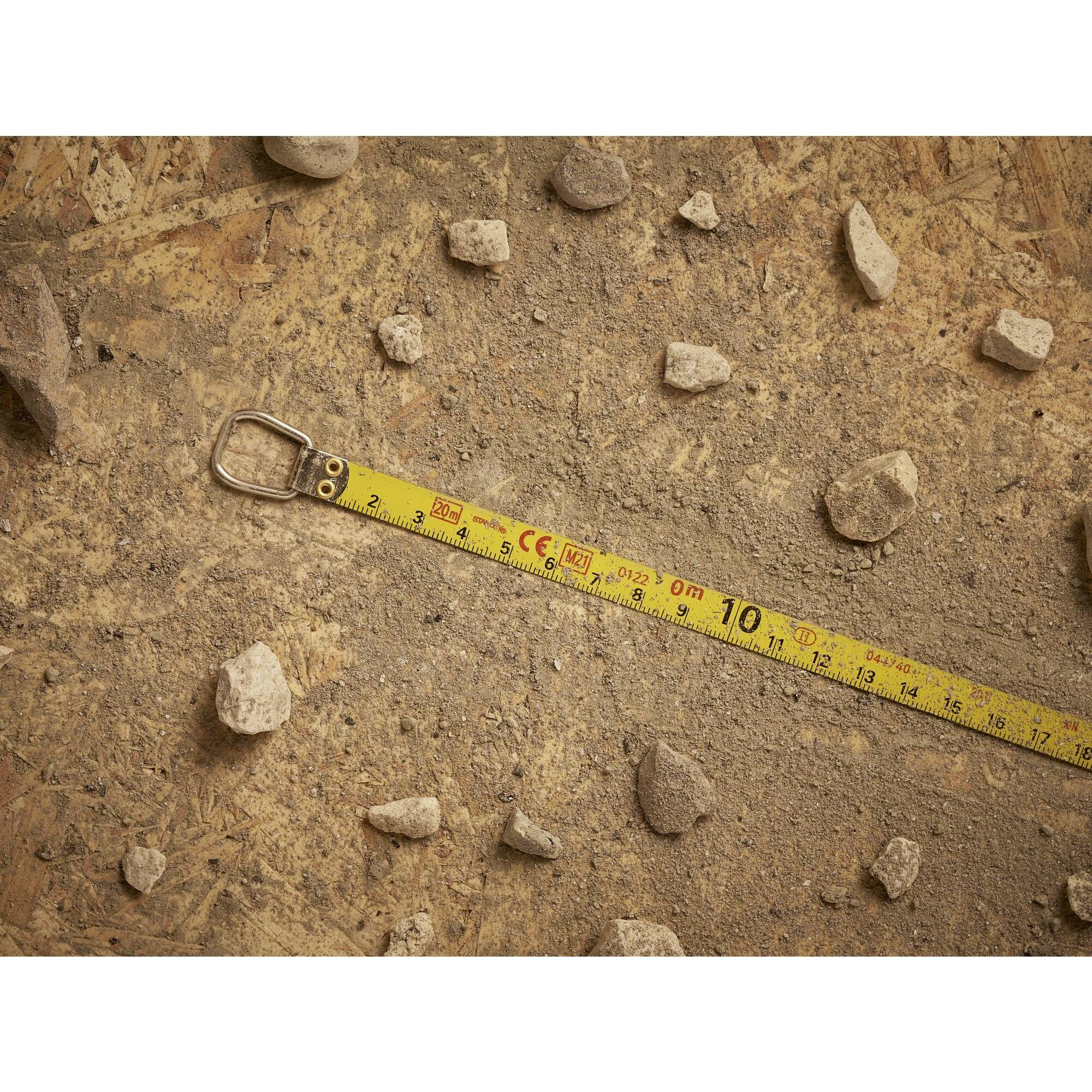 A yellow tape measure lies coiled on an earthy surface, surrounded by small stones.