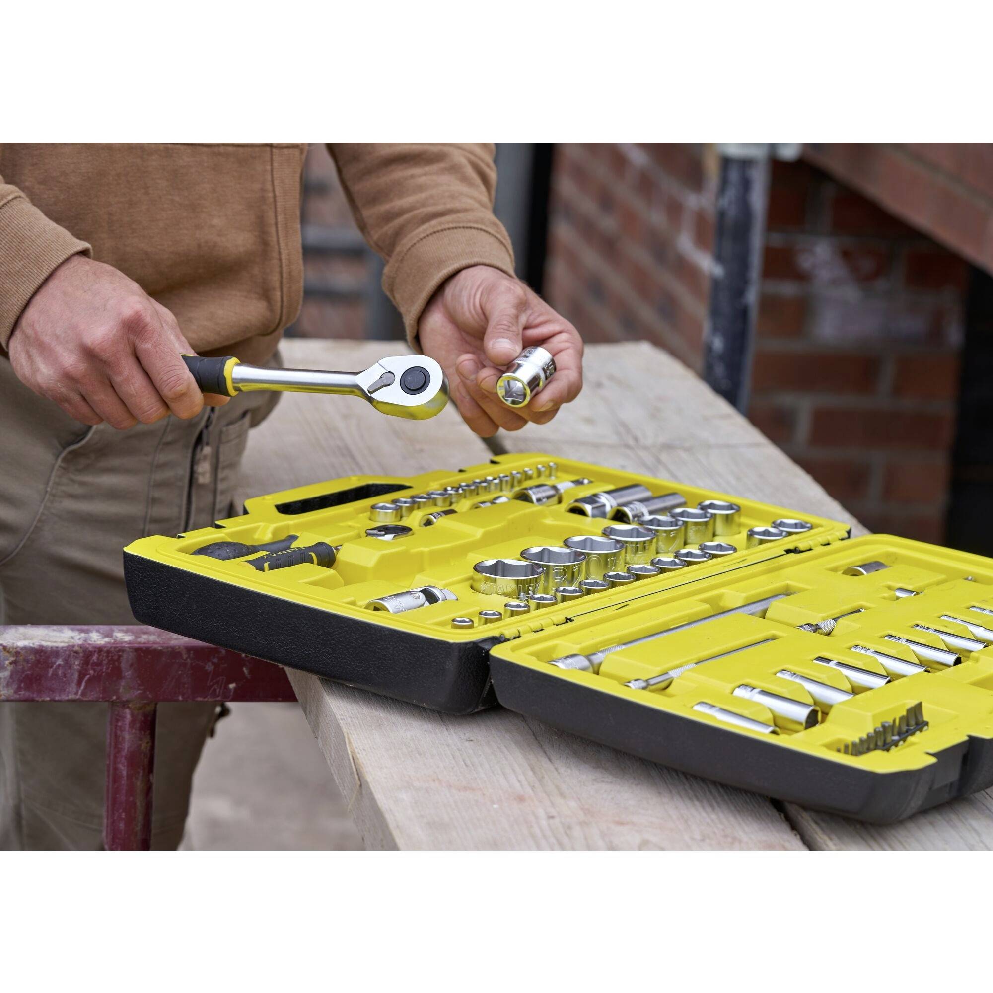 A person is holding a spanner and a screw whilst standing in front of a yellow toolbox resting on a table.