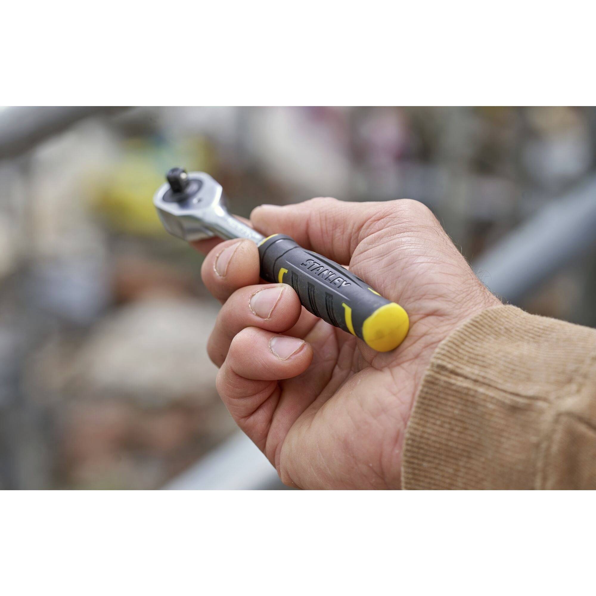 A hand holds a ratchet spanner against a blurred background. The handle of the spanner is black and yellow.