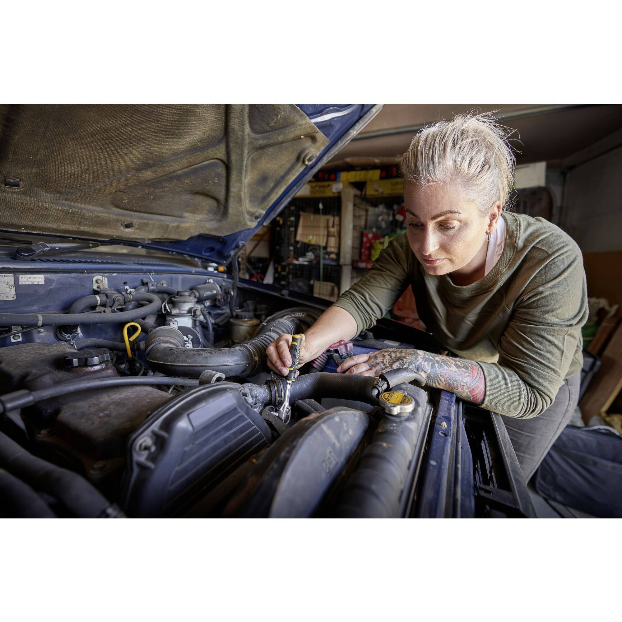 A woman with tattoos is repairing an engine in a workshop, focused on her work while holding a tool.