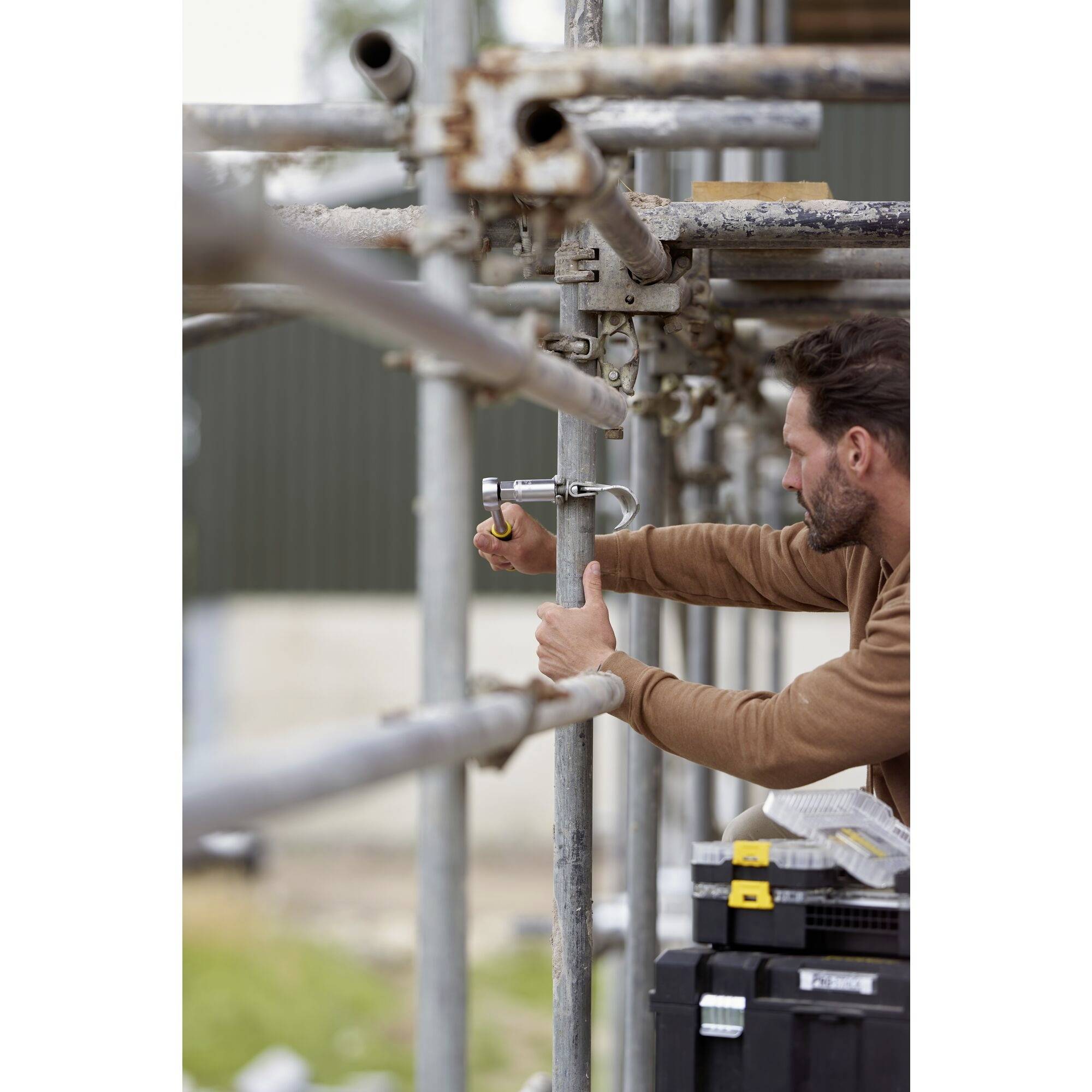 A man is repairing a scaffold outdoors. He is using a tool to tighten a connection. Construction elements can be seen in the background.
