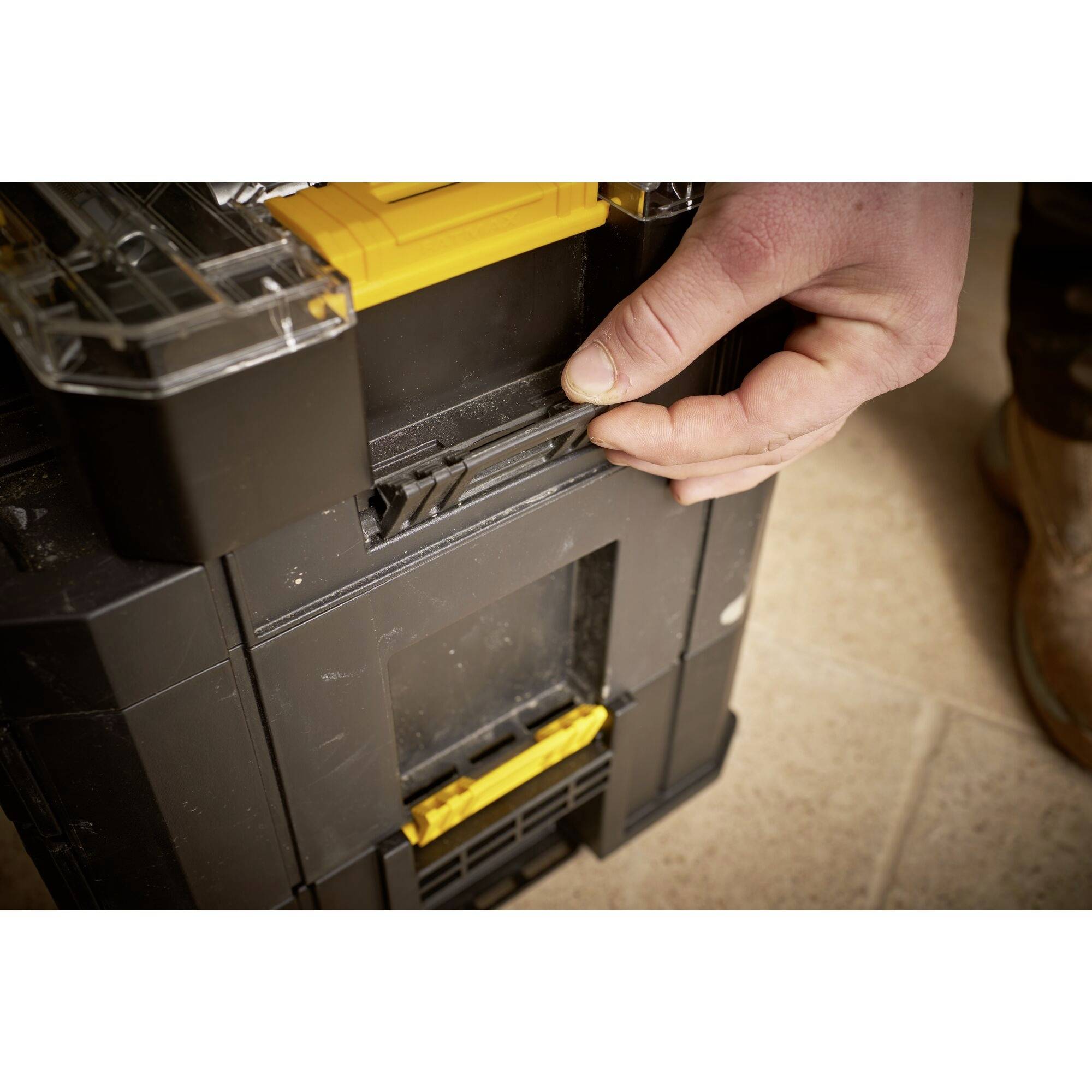 A person opens a black toolbox with a yellow fastener by hand on a beige floor.
