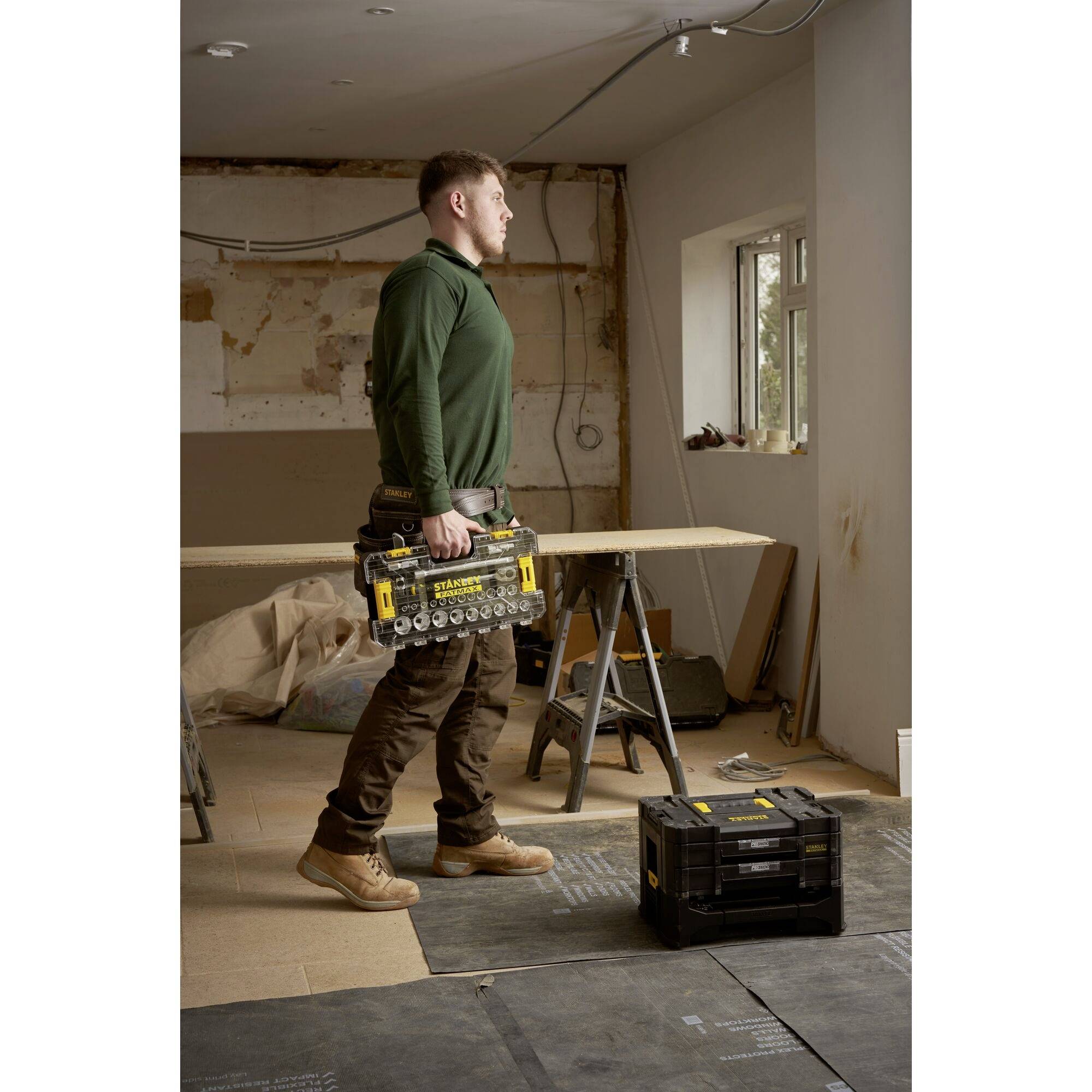 A man is carrying a toolbox in an unfinished workshop with windows. Sawhorses and tools are visible in the background.