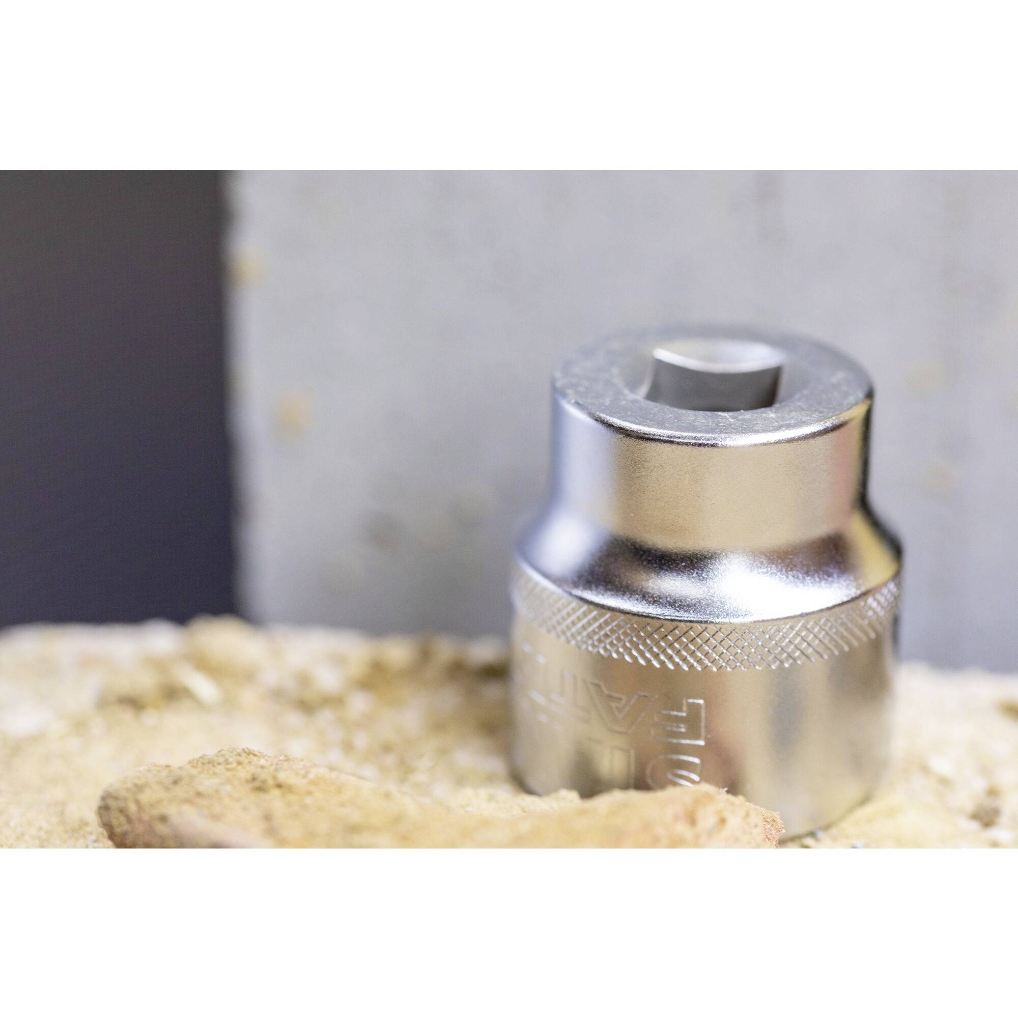 Metal socket wrench head resting on a sandy surface against a grey background. Tool gleams in the light, with a simple, functional design.