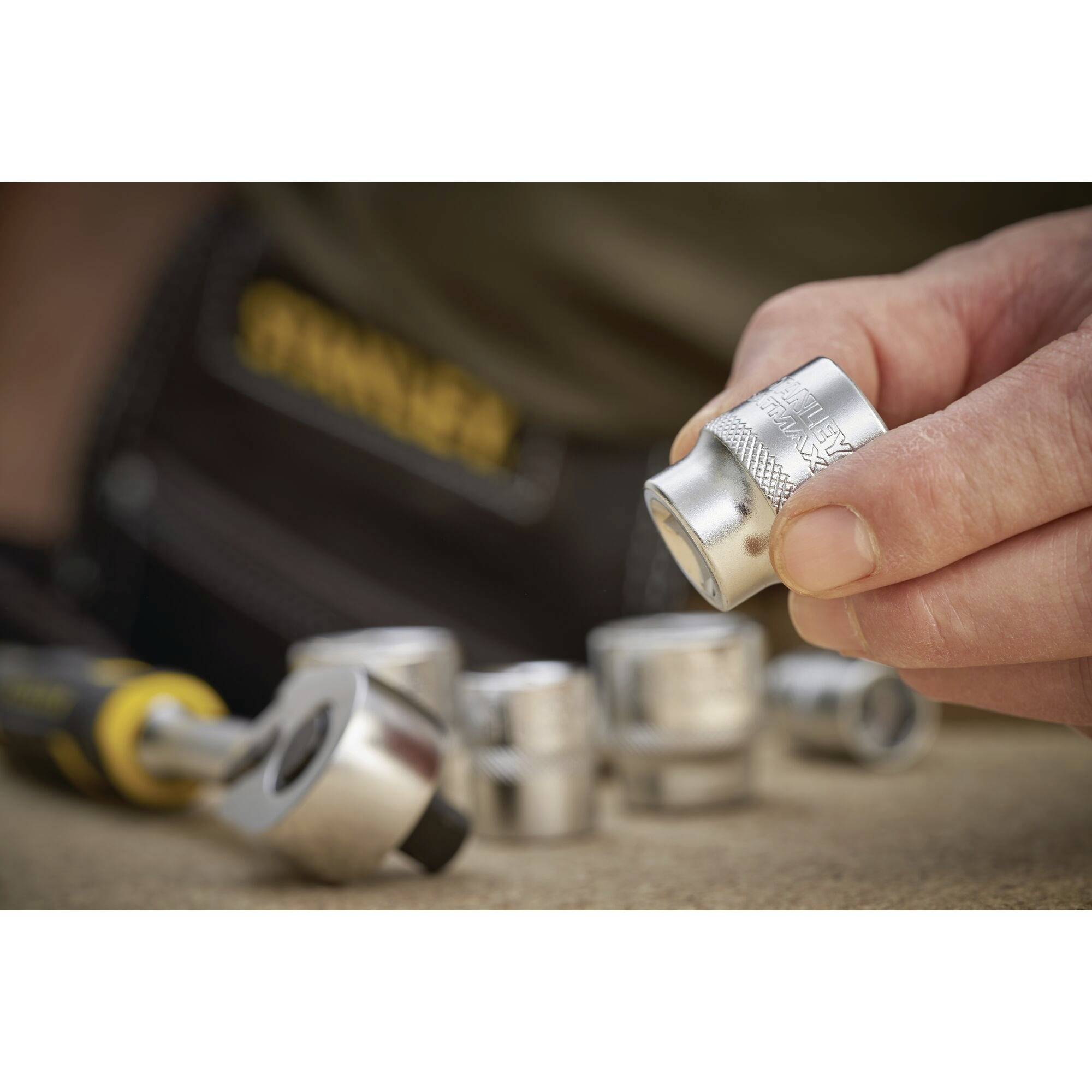 A hand holds a silver stud bolt in front of other stud bolts and a tool, which are blurred in the background.