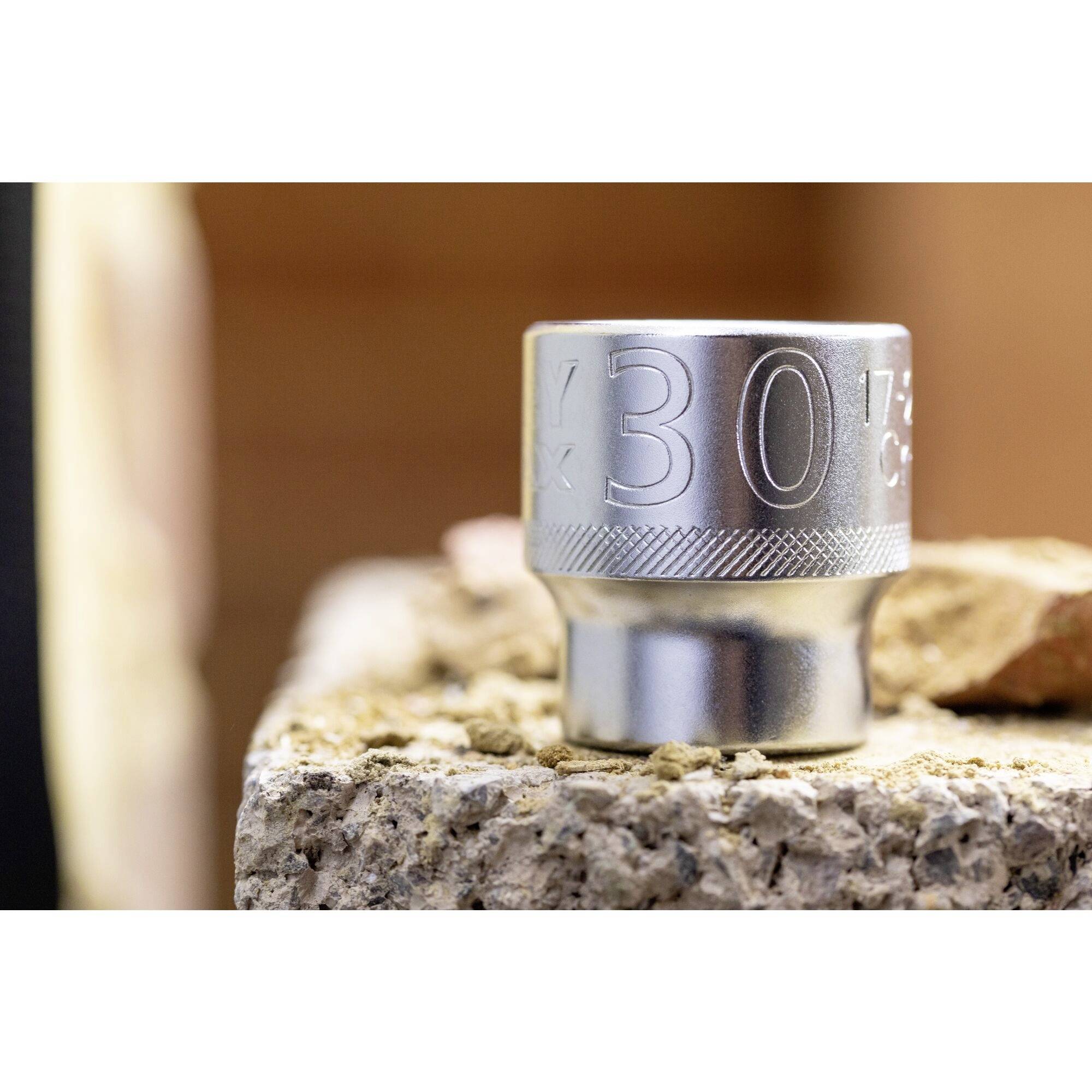 A silver 30-mm socket lies on a concrete block. Warm, blurred colour tones are visible in the background.